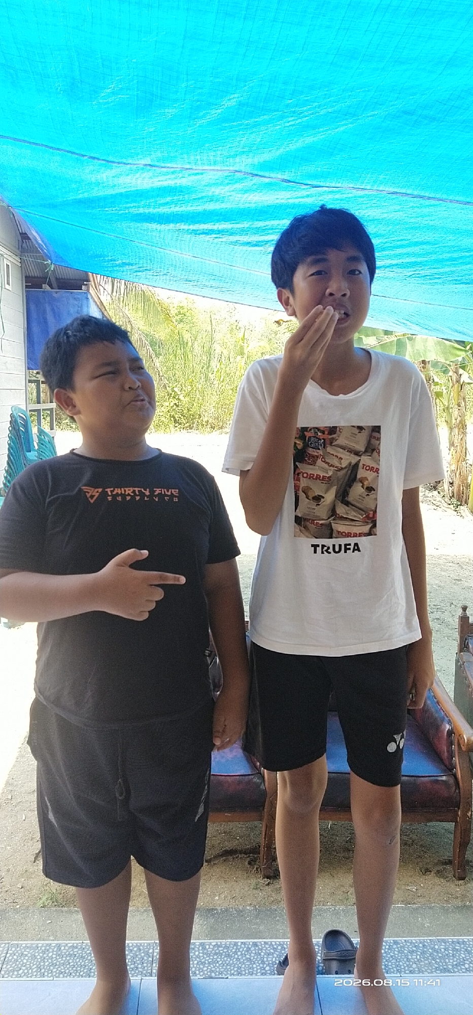 Two young boys are standing outdoors under a blue tarp. The boy on the left, wearing a black t-shirt and black shorts, is pointing his left index finger towards the viewer and looking to his left with a grimacing expression. The boy on the right, wearing a white t-shirt with a print of potato chip bags and black shorts, has his right hand near his mouth, with his mouth open and teeth showing. The background consists of dry vegetation and a building. A vintage-style chair is partially visible to the right. The scene appears to be in Muka Haji, Indonesia.