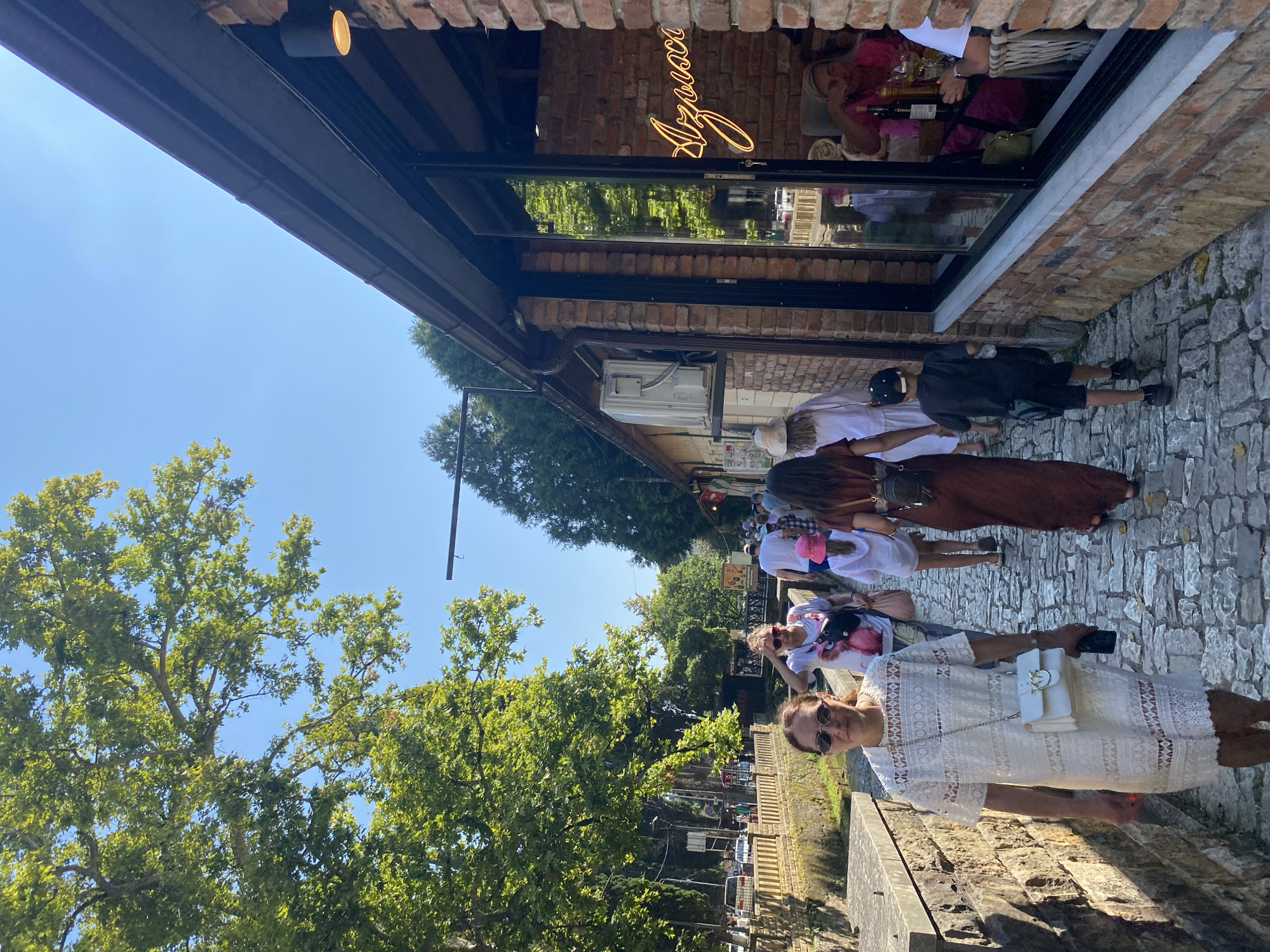 The image shows a cobblestone path lined with a low stone wall on the left and a brick building with large windows on the right. A group of people are walking along the path on a sunny day. The building has a neon sign in the window that says "Azovsc". A few people are seated inside the building, some of whom appear to be eating or drinking. Trees with green foliage overhang the path and the building. The sky is clear and blue.