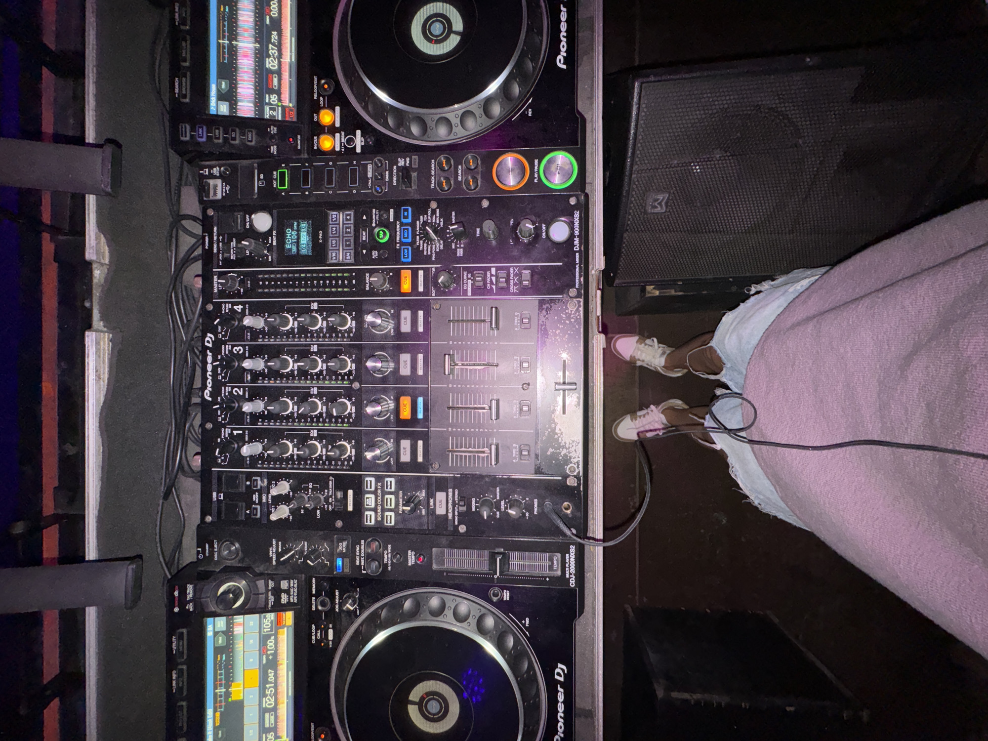 The scene is an indoor entertainment venue, such as a nightclub or bar, described as being in Moscow, Russia. The image is captured from a first-person perspective, looking down at a professional DJ setup and the lower body of an individual operating it.
The central focus is a black DJ console comprising two Pioneer CDJ-2000NXS2 multi-players flanking a central Pioneer DJM-900NXS2 professional mixer. The left multi-player displays a waveform with green and yellow segments, showing a time of 02:51.047 and a tempo of 105 BPM. Its jog wheel is illuminated blue, and the 'Play/Pause' button is glowing orange. The right multi-player also displays a waveform, indicating a time of 02:37.724 and a tempo of 105 BPM. Its jog wheel is illuminated green, and the 'Play/Pause' button is glowing green.
The central mixer features four channels with various knobs, faders, and illuminated buttons, including controls for Sound Color FX and Beat FX. The 'ECHO' Beat FX is active, displaying "105 BPM" in blue. Numerous black cables connect the components of the DJ setup. The equipment rests on a dark, flat surface. Above the console, a dark, textured wall or partition is visible, with additional cables draped across it.
The lower half of a person is visible at the bottom of the frame, standing behind the console. They are wearing light blue ripped jeans and white sneakers with pink accents. A pink or purple blanket or shawl covers their upper legs. A thin black cable extends from their feet down to the dark floor.
To the right of the person's feet, a large, black, rectangular speaker with a metallic grille and a prominent white "W" logo is visible on the floor. A dark, box-like object, possibly another speaker or equipment case, is partially visible to the left. The overall lighting is dim and ambient, with purple and orange hues cast upon various controls and surfaces of the DJ equipment, indicating an active club environment.