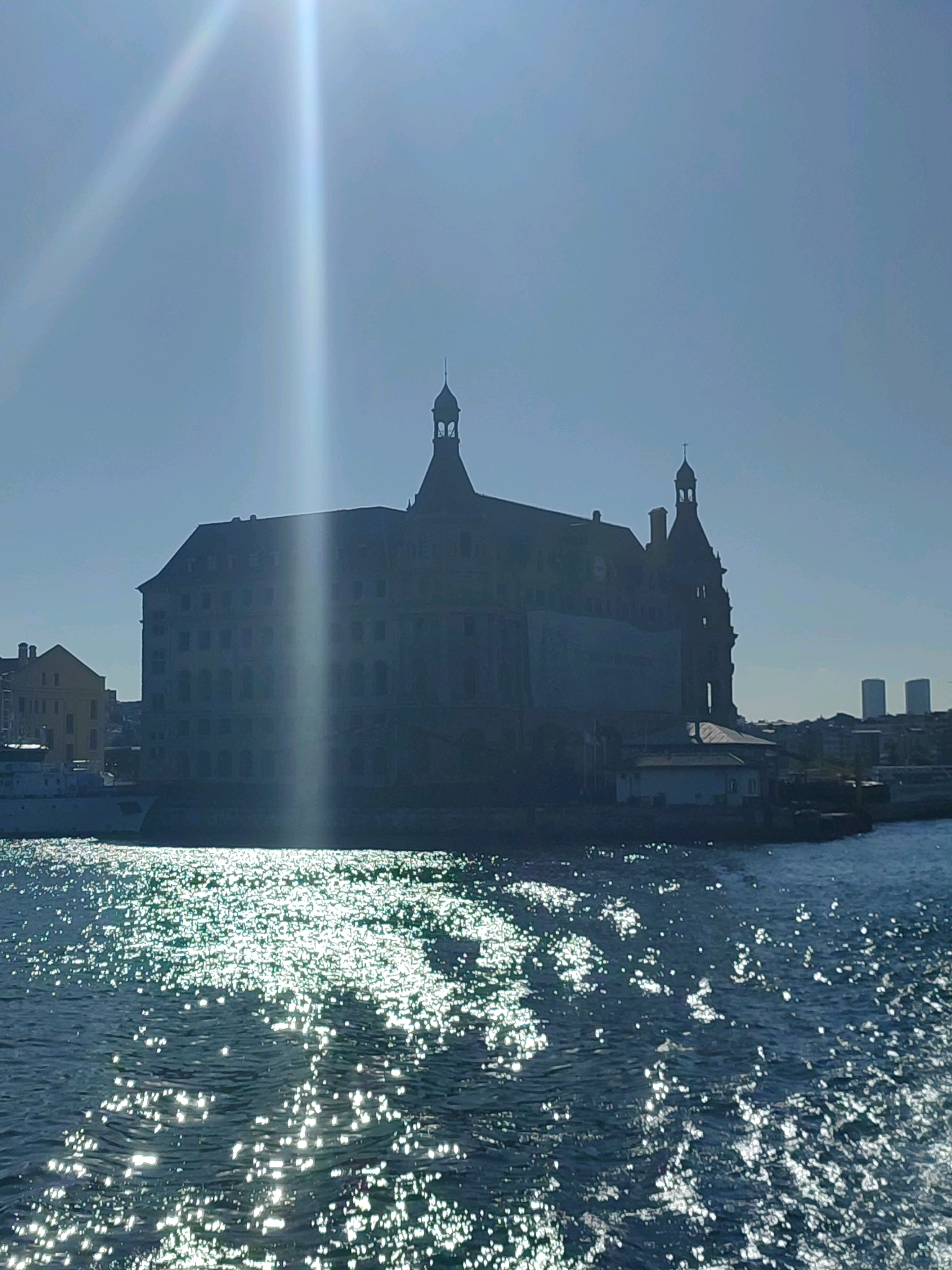 The image shows a wide shot of the Haydarpaşa Train Station in Kadıköy, Istanbul, Turkey, from a water perspective. The sun is bright, casting a strong glare across the sky and reflecting off the water in the foreground, creating a shimmering effect. The train station, a large, historic building with distinctive towers and architectural details, is silhouetted against the bright sky. To the left of the station, another building and a white boat are partially visible. In the distance, on the right side of the frame, two modern skyscrapers stand out against the horizon, indicating the urban landscape of Istanbul. The water in the foreground is choppy, with numerous light reflections scattered across its surface. There are no people visible in the image.