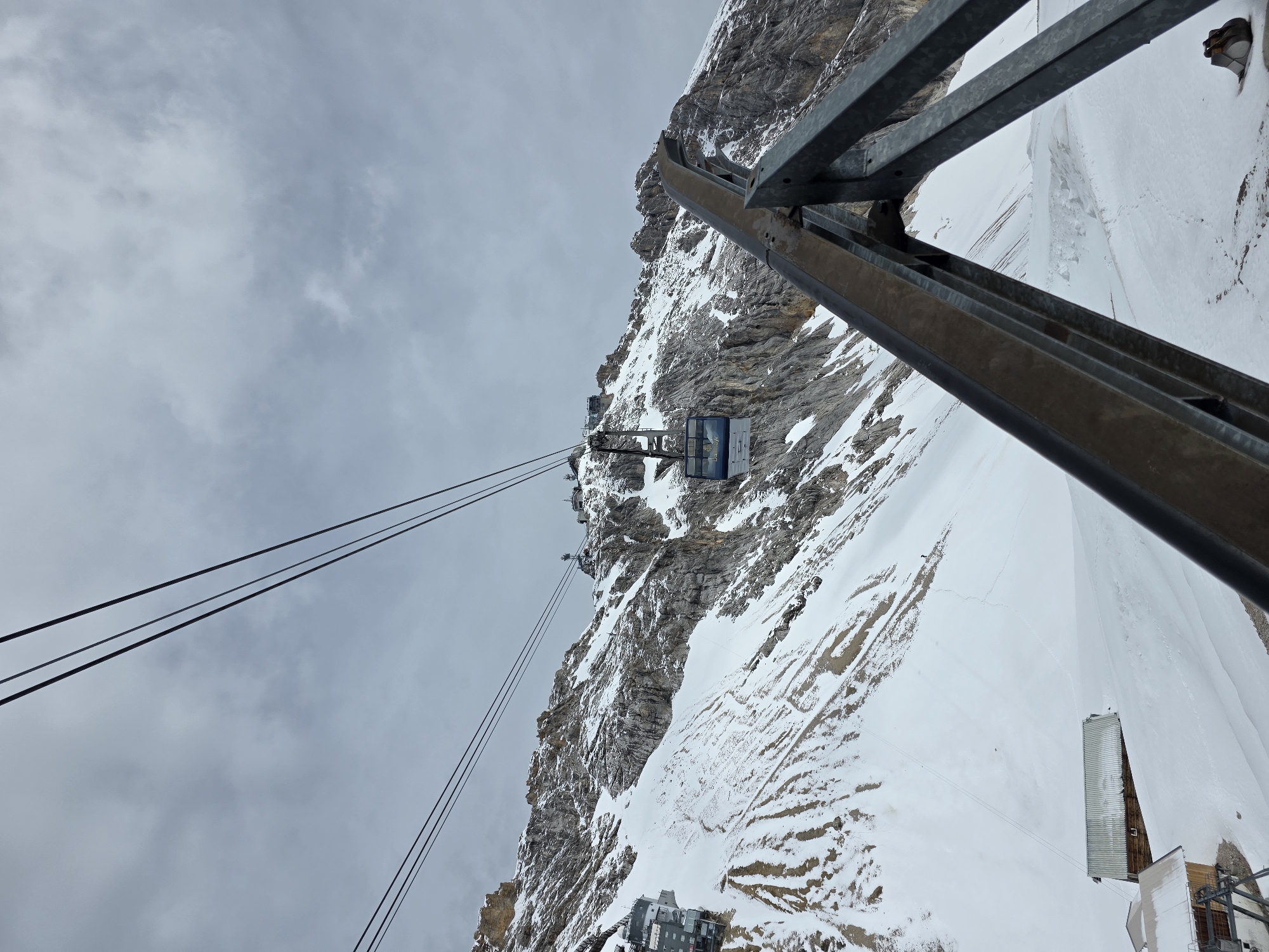 This image captures an alpine scene featuring a cable car system ascending a snow-covered mountain on an overcast day in the Dachstein region, Austria. The sky is uniformly light grey and cloudy.
In the foreground, a large, dark metal support structure, likely part of a cable car pylon or station, dominates the lower right, composed of thick, angled beams. Multiple parallel steel cables extend from the lower left and ascend into the upper right, receding towards the mountain summit. A blue and white cable car cabin, with "Dachstein" prominently displayed on its side, is visible in the mid-ground, suspended from these cables and traveling uphill.
The mountain itself is steep and rugged, characterized by extensive snow cover interspersed with exposed sections of grey and brown rock. The snow shows varying textures, with some areas exhibiting linear patterns from natural accumulation or past activity, and darker patches where rock or earth show through. Higher on the mountain, near the presumed upper station, several modern-looking buildings with grey facades are visible, integrated into the rocky terrain.
At the base of the mountain, in the lower left, are additional functional structures, possibly part of a lower cable car station or maintenance facility. Further down and to the left, two smaller, rustic wooden buildings with corrugated metal roofs are situated on a snow-covered plain. Footprints or tracks are visible in the snow around these structures. In the far lower right, partially obscured by the foreground metal structure, a yellow excavator bucket is visible resting in the snow. No individuals are discernible in the scene.