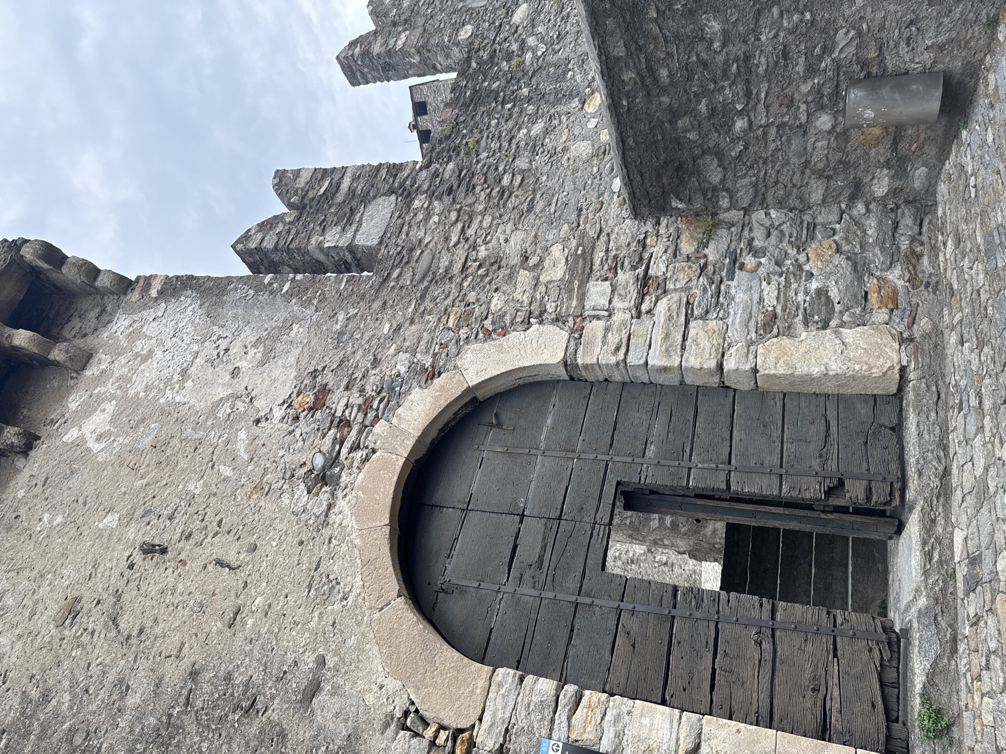 The image shows a portion of the Castel Grande in Bellinzona, Switzerland. The foreground features a large, imposing wooden double door, with one leaf ajar, revealing a set of stone steps leading upwards. The door is reinforced with metal bands and studs. The archway above the door is constructed of large, rough-hewn stones. The surrounding walls are made of weathered, grey stone, with visible mortar lines and varying sizes of irregular stones. To the right of the door, a section of the castle wall continues upwards, featuring battlements against a cloudy sky. Some greenery is visible growing from cracks in the stonework. In the lower right corner, a modern, cylindrical metal trash can is mounted on the wall. The ground is paved with cobblestones.