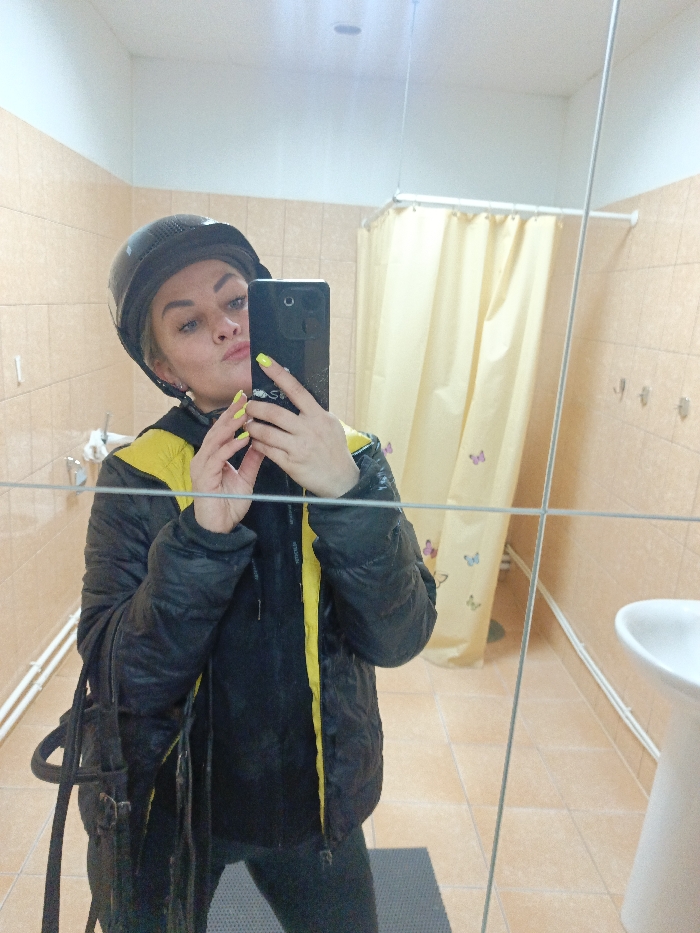 In a restroom facility in Gorka, Russia, a female individual takes a mirror selfie. She wears a black riding helmet and a black puffer jacket with yellow interior lining. Her fingernails are painted neon yellow, and her lips are pursed. She holds a dark-colored smartphone, partially obscuring her face. A dark strap, consistent with a horse bridle or bag, hangs from her left shoulder. The background features light beige tiled walls, a white pedestal sink partially visible on the right, and a yellow shower curtain with multi-colored butterfly decals hanging from a white rail. White heating pipes are visible along the lower left wall. The floor is covered in beige tiles with a dark mat in the foreground.