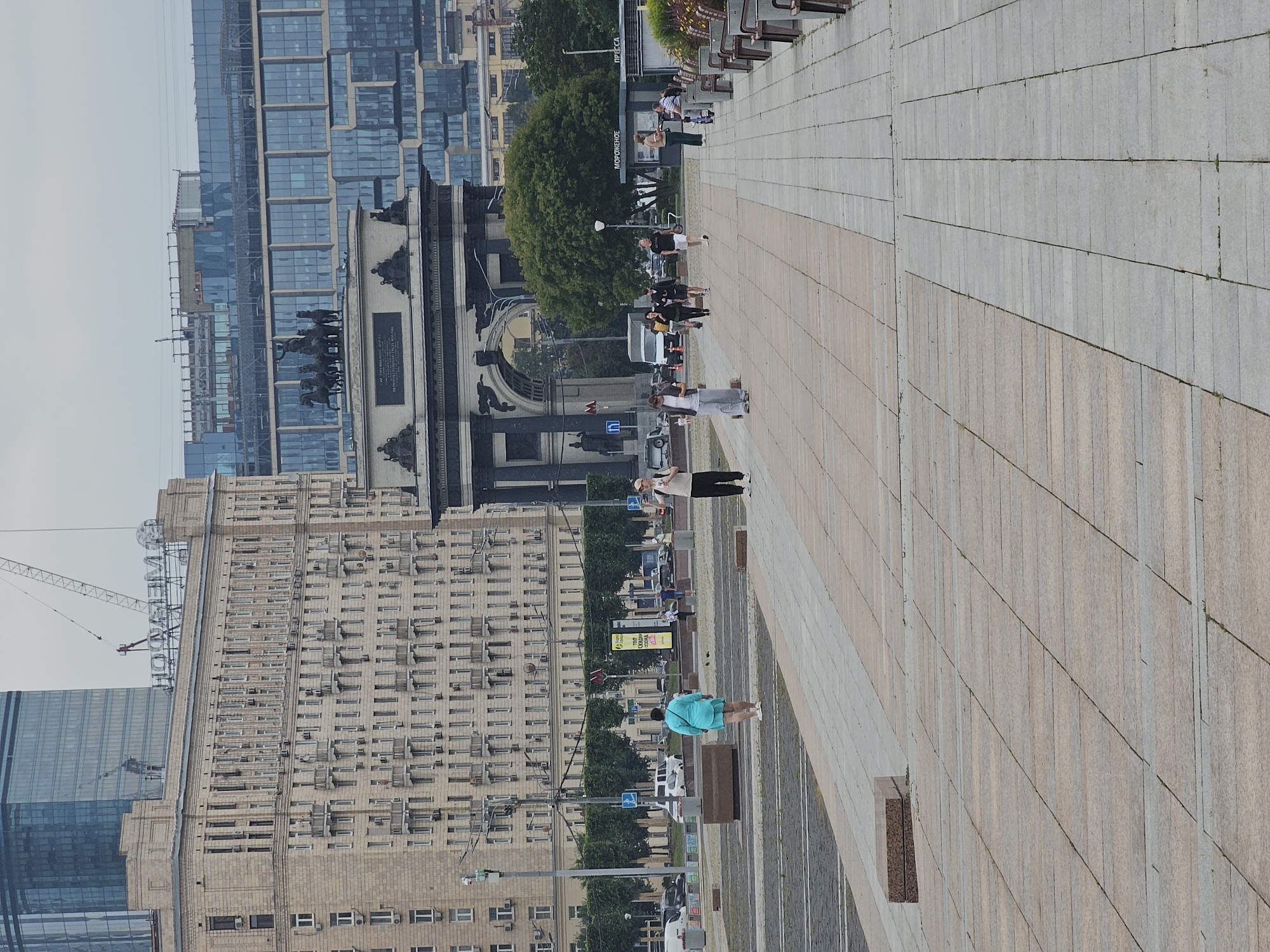 The image depicts a wide pedestrian walkway in Moscow, Russia, featuring a prominent triumphal arch in the background. The walkway is paved with large, light-colored stone tiles arranged in a pattern, with a band of darker stone tiles bordering the central area.
Several people are visible walking along the pathway. In the mid-ground, a woman in a bright turquoise dress is walking away from the camera. To her right, a group of individuals, some in casual attire, are walking towards the arch. Further in the distance, other pedestrians can be seen, as well as a kiosk with the word "МОРОЖЕНОЕ" (ice cream) and "ПРЕССА" (press) written on it.
Dominating the background is a grand triumphal arch, adorned with sculptures and inscriptions, with a quadriga atop its central structure. Behind the arch, a modern glass-fronted building stands tall, reflecting the sky. To the left of the arch, a large, older building with a pale facade and numerous windows and balconies lines the street. Trees are planted along the perimeter of the walkway, and streetlights and traffic signs are also visible. The overall scene suggests a public square or avenue in an urban environment, likely a well-known landmark.