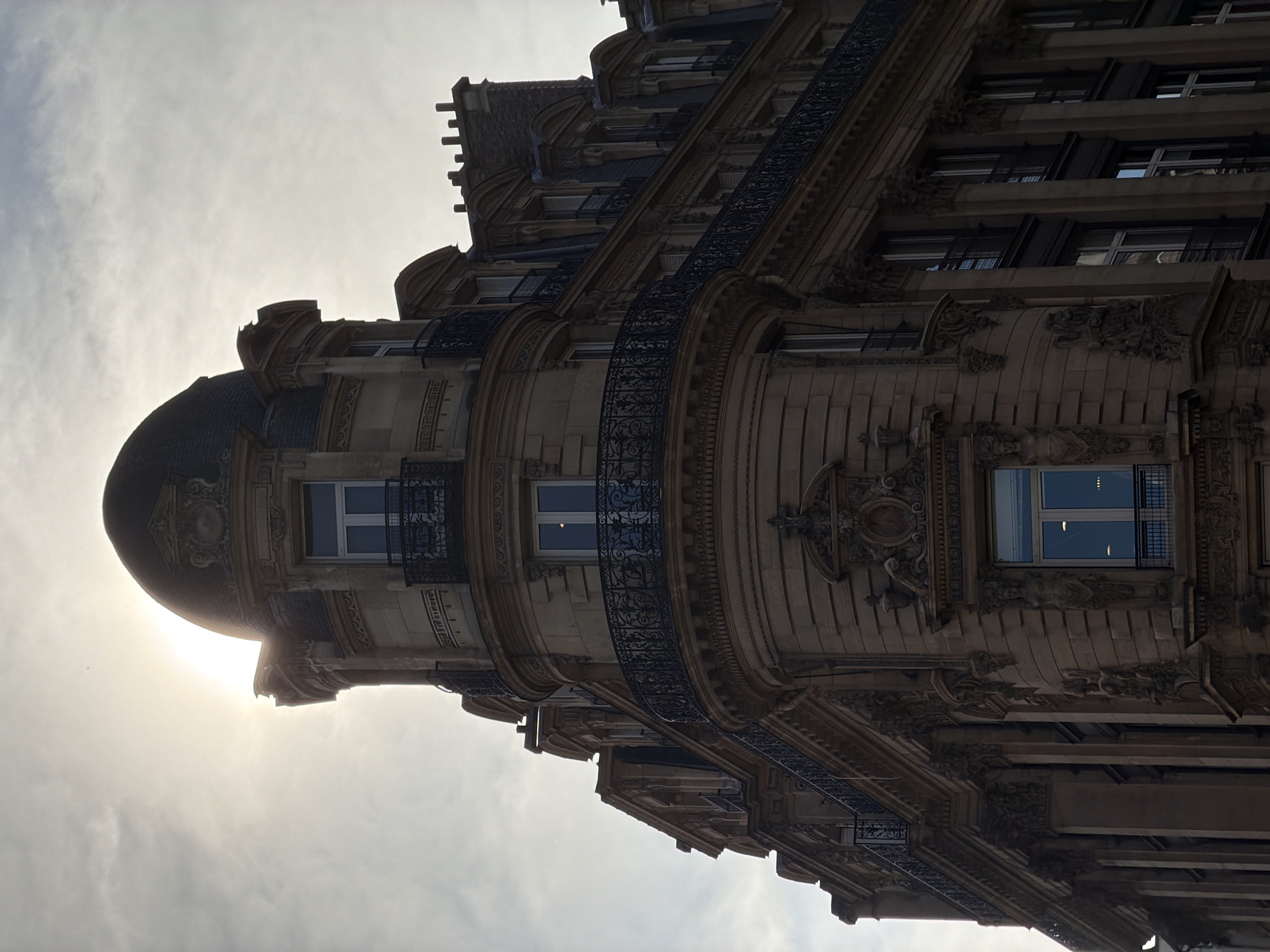 An ornate, multi-story building, characteristic of Parisian architecture, dominates the frame. The image is taken from a low angle, looking up at the facade against a bright, cloudy sky. The building features a prominent curved corner with a dark, domed turret at the top. The sun's glow is visible from behind and to the left of the dome, suggesting either early morning or late afternoon.
The facade is richly detailed with decorative stone carvings, cornices, and pilasters. Multiple windows, some recessed, are visible across the building's levels. Elaborate black wrought-iron balconies with intricate scrollwork extend along the curved section and flatter parts of the building. The windows reflect the bright sky and interior lights in some panes. No specific activity or people are visible, focusing solely on the architectural grandeur. The overall impression is one of classic urban elegance in Paris, France. There is no visible text in the image.
