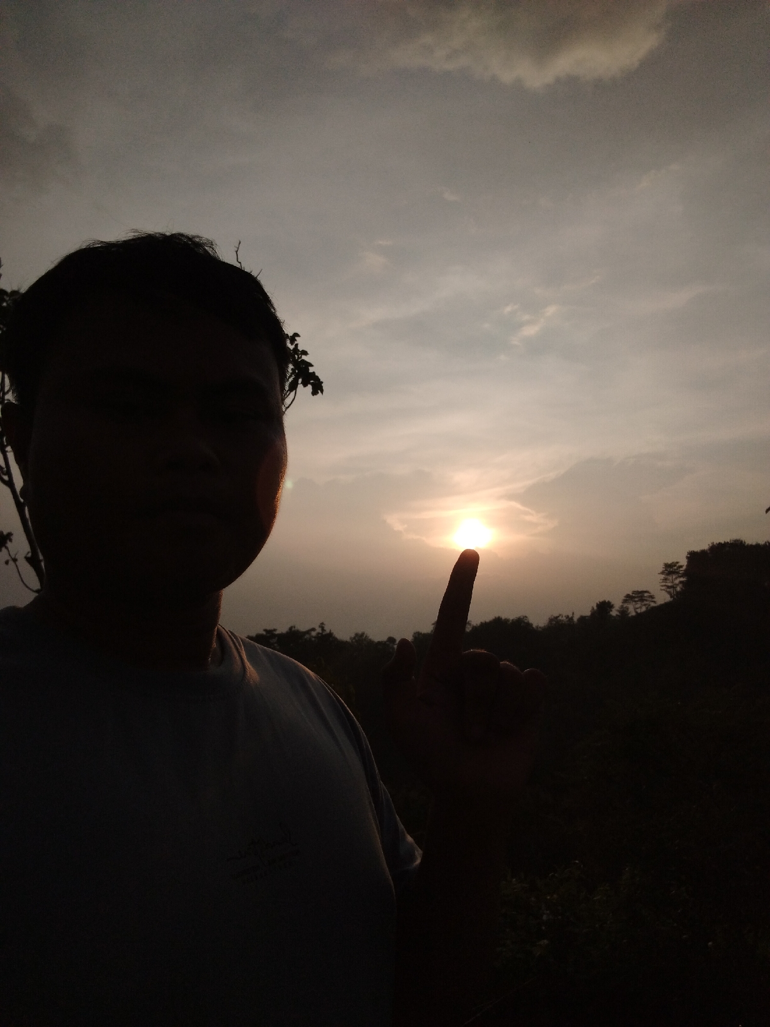 A silhouette of a person is positioned in the foreground of the image, with their index finger pointing towards the setting sun, which is slightly obscured by clouds. The background features a landscape of trees and a cloudy sky. The location appears to be a natural setting, possibly a rural or semi-rural area. The city is not identifiable from the image.