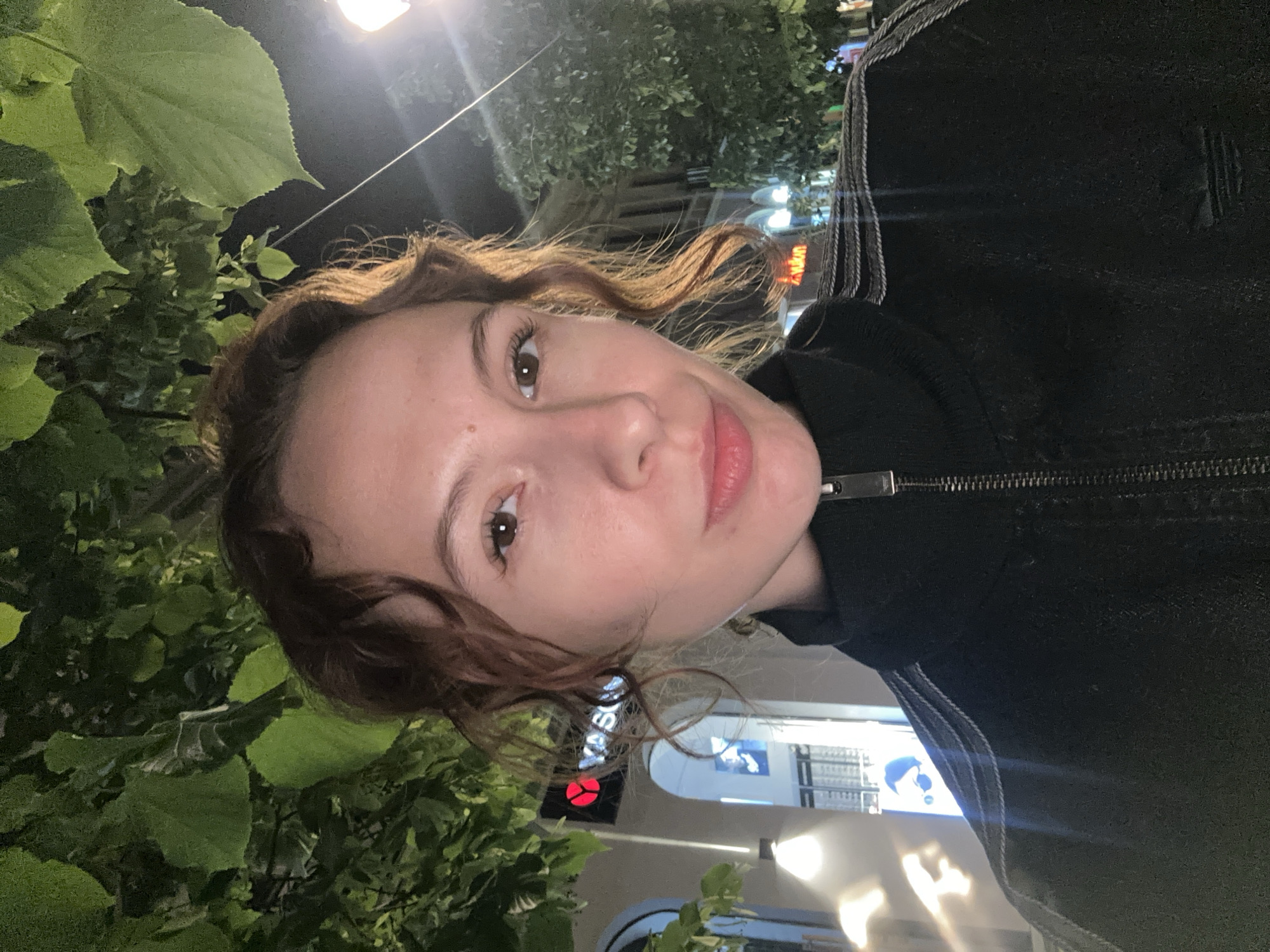 A young woman is captured in a close-up, illuminated selfie taken outdoors at night. Her face, framed by light brown wavy hair, is central, with a calm and natural expression. Her right eye is prominent, looking towards the camera, and her lips have a soft pink hue. She is wearing a dark, likely black, zip-up jacket or hoodie, identifiable by a metallic zipper pull and a subtle textured or striped detail along the arm/side.
The background is dark, indicative of nighttime, with artificial lights from streetlamps or buildings providing illumination. Lush green foliage, likely from trees or bushes, is visible in the upper left and lower right corners, suggesting an urban or park-like setting. In the distance, blurred lights and parts of buildings can be seen. Notably, a storefront sign with a red circular logo and partial text "ASON" is visible behind her left shoulder. Another blurred sign in the deeper background shows hints of Latin script ("yukun"). The scene is set in an urban environment in Novi Sad, Serbia.