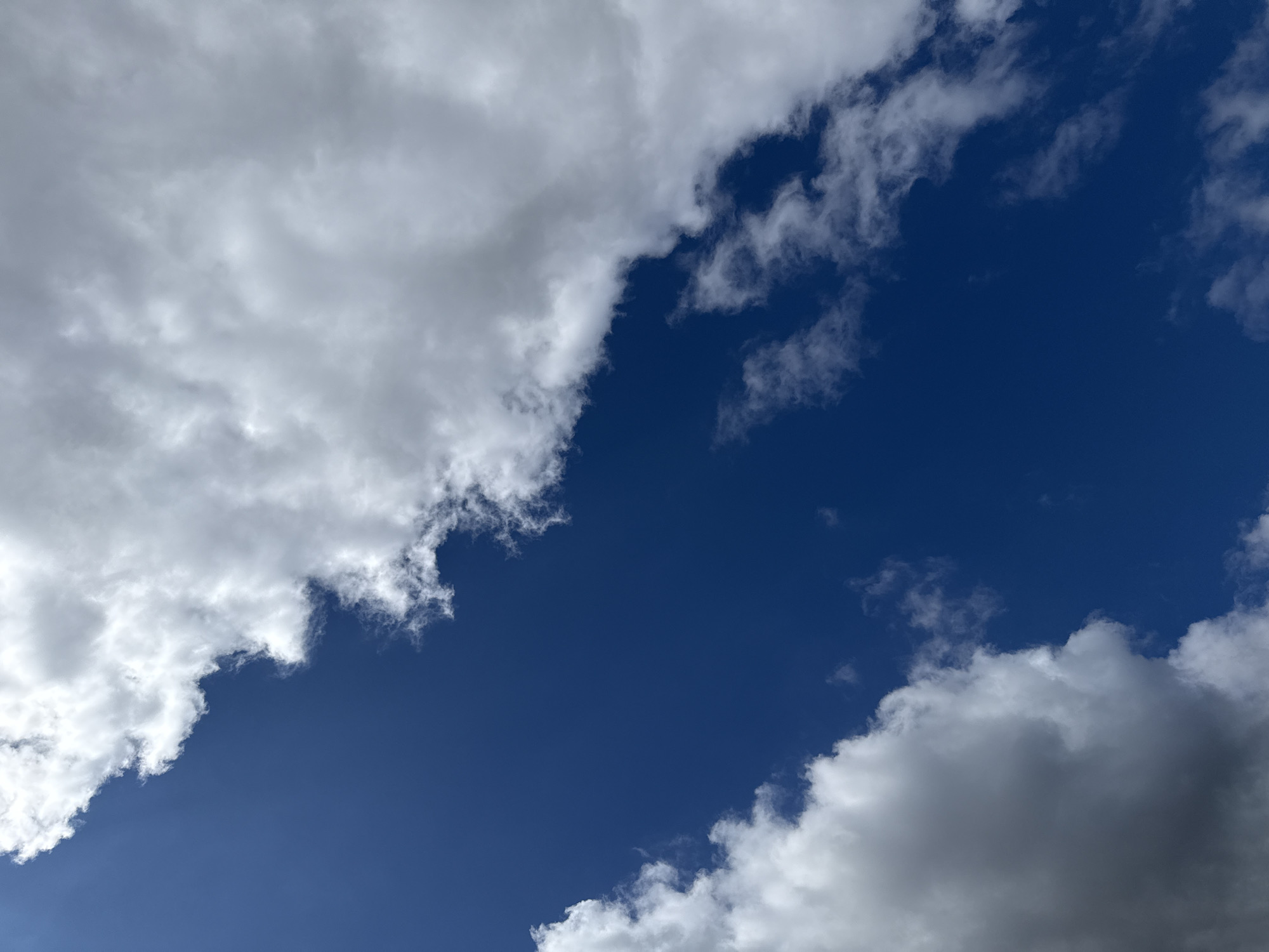 The image is a low-angle shot of a deep blue sky partially obscured by white and grey clouds. The clouds are textured and appear fluffy in some areas, with darker, more ominous patches in others. The sky is clear and vast, indicating an outdoor setting. The image appears to have been taken in Großpösna, Germany.