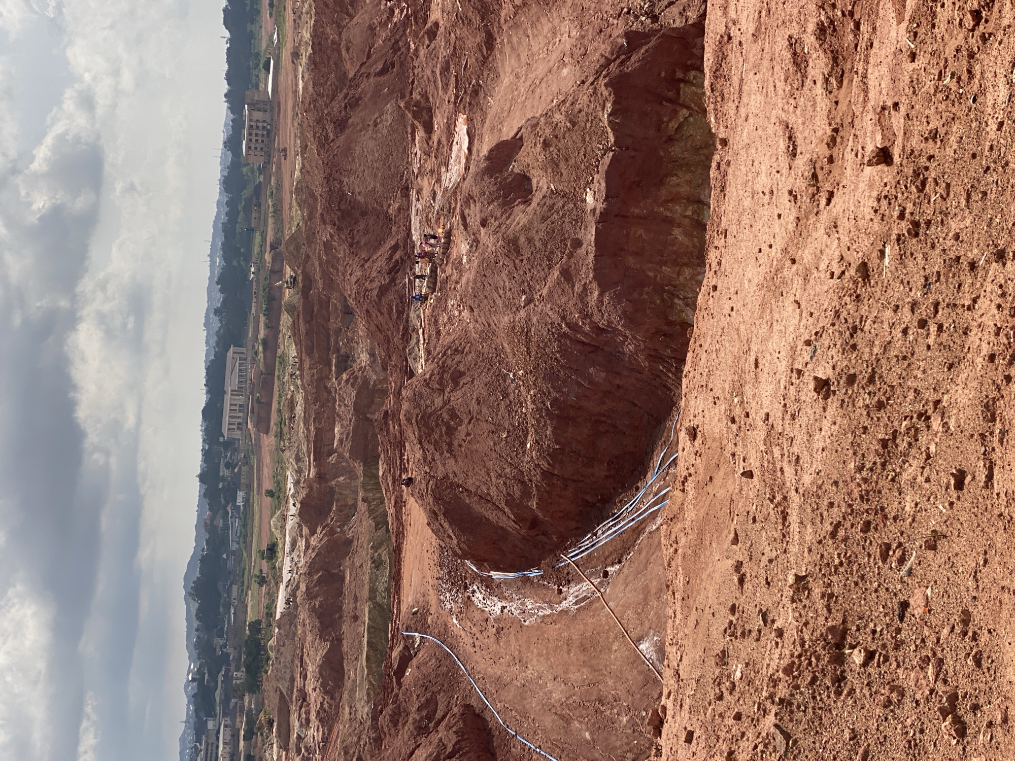 The image depicts an active construction or excavation site in Reifild-Ist, Nigeria. The scene is characterized by a large, reddish-brown earth embankment, likely a man-made cut into the terrain. The sun appears to be high in the sky, casting strong shadows and suggesting it is daytime. The weather appears fair with some scattered clouds visible on the left side of the frame.
Several people are visible on the embankment, dressed in casual clothing. Some appear to be working, possibly overseeing or participating in the excavation. In the mid-ground, there are several blue pipes laid out on the ground, suggesting water or drainage infrastructure is being installed. A large, excavated pit is the focal point of the activity, with machinery or vehicles possibly present but not clearly identifiable.
In the background, a semi-urban or rural landscape unfolds, featuring buildings and vegetation. The architecture of the buildings suggests a residential or commercial area. The overall impression is one of significant human activity related to construction and development, set against a natural, earthy backdrop. No visible text is present in the image.
