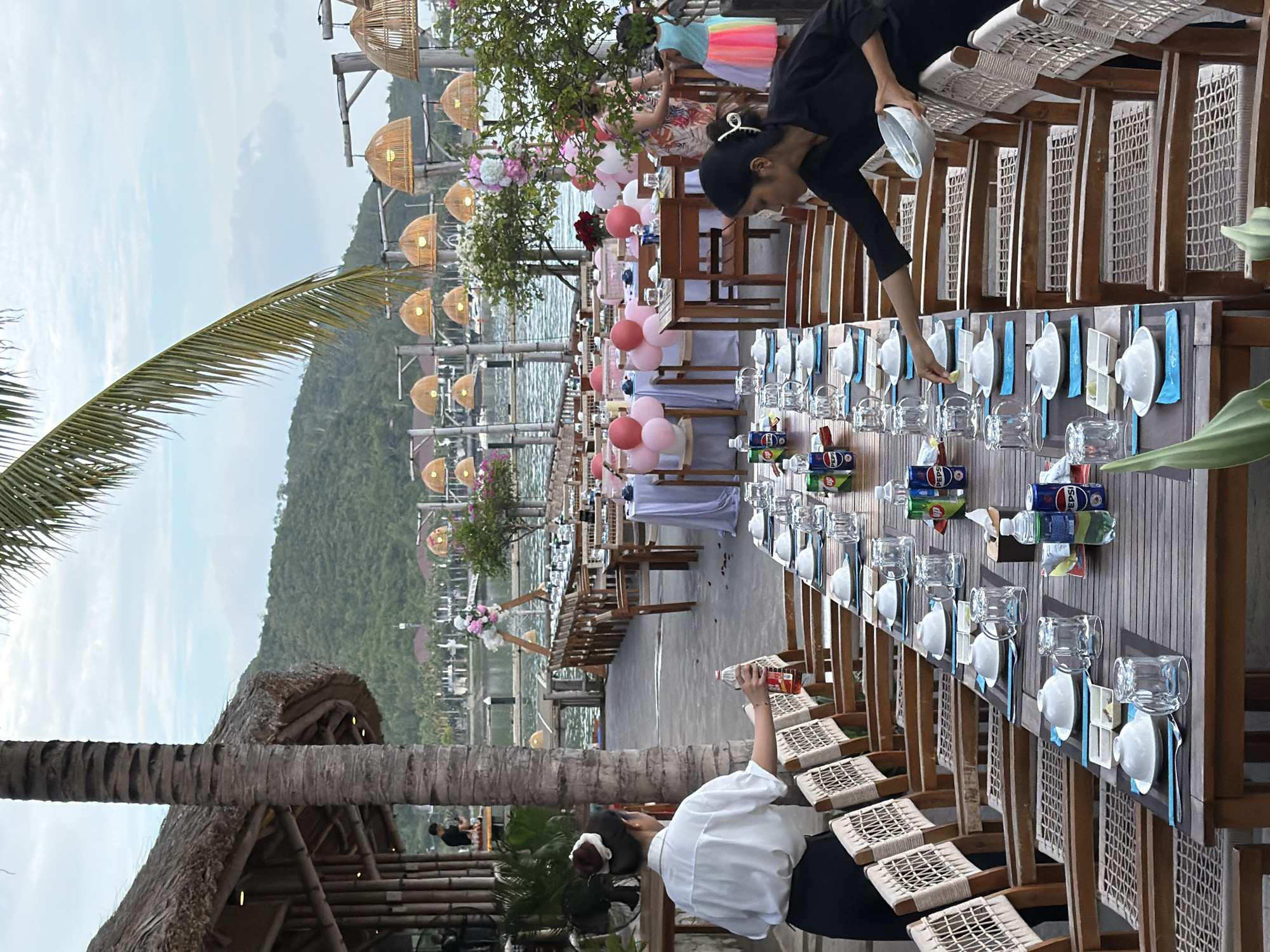 The image depicts an outdoor dining setup by a body of water, likely in Nha Trang, Vietnam, with a scenic backdrop of lush green hills. In the foreground, a long, wooden banquet table is meticulously set for a meal, adorned with white plates, bowls, glasses, and blue placemats. Canned beverages, including Pepsi, and bottled water are also present on the table. Two waitstaff members are visible; one, a woman in a white shirt and black pants, is standing near the left side of the table, holding a bottle of liquid. The other, in a black shirt and black pants, is in the process of placing a white bowl onto the table setting. Behind the main table, more tables are arranged, some draped with white tablecloths and decorated with pink and red balloons, suggesting a festive occasion. In the background, a thatched-roof structure is partially visible, and a line of hanging lamps made of woven material illuminates the area. The water reflects the sky and the distant shoreline, which is dotted with buildings and vegetation. A young girl in a brightly colored skirt can be seen near the tables in the middle ground. The overall atmosphere is one of a prepared outdoor event, possibly a restaurant or a venue hosting a party.