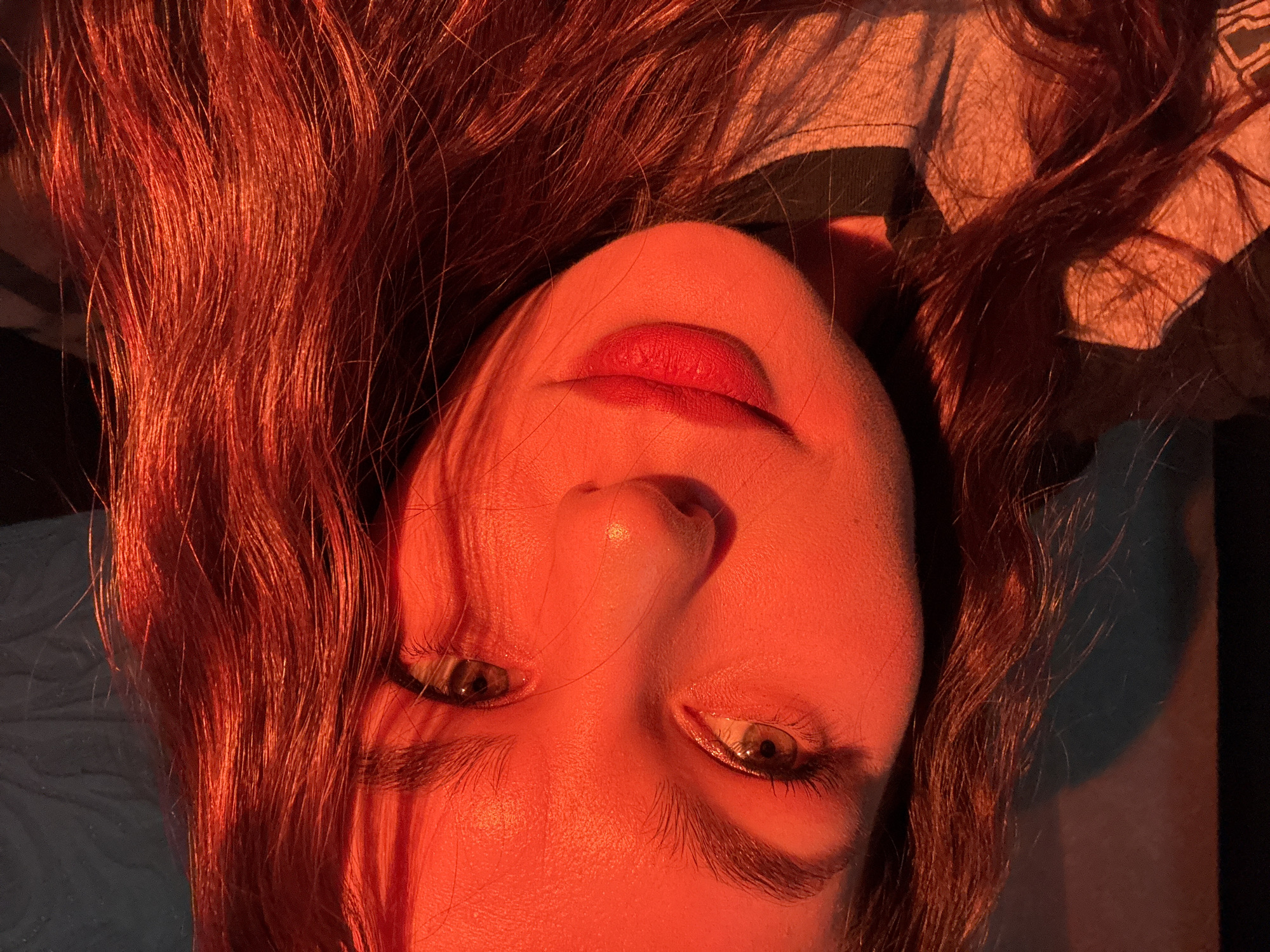 A close-up, slightly rotated portrait of a woman with red hair bathed in warm, orange light. Her face is tilted upwards, eyes looking towards the viewer. She has full red lips, defined eyebrows, and long eyelashes. The hair is wavy and voluminous, catching the light and creating highlights. The background is partially obscured by the hair and shadow, revealing hints of clothing. It is impossible to verify the exact location as Chaikovsky, Russia, from the image.