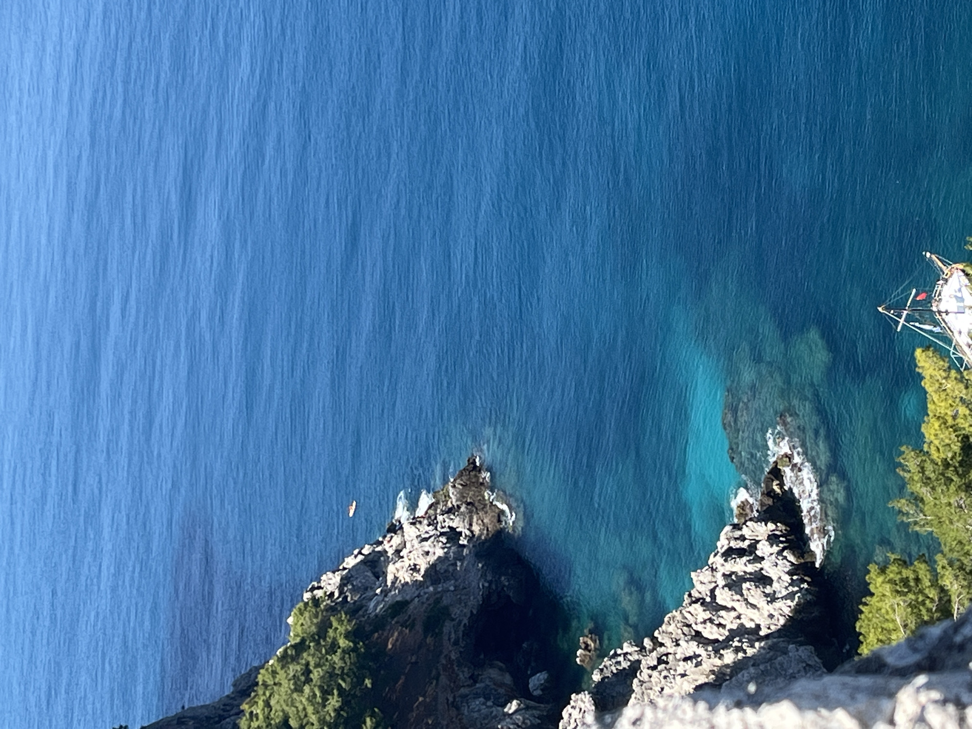 An aerial view shows a scene in Alanya, Turkey. The deep blue sea takes up most of the frame. Jagged, rocky cliffs covered with trees extend into the water. A small, isolated rock formation with a boat is visible in the water. A kayak is seen near another set of rocks. A sailboat is next to the trees on the right of the image.
