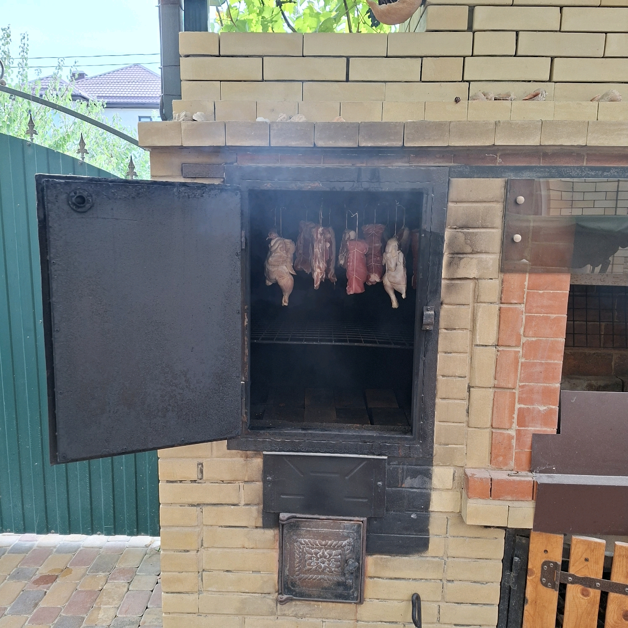 An outdoor brick oven with a dark metal door ajar is shown in Anapskaya, Russia. Inside the oven, several pieces of meat and two whole chickens are hanging from hooks. The meat appears to be raw and is tied with white string. Smoke is visible inside the oven, indicating that it is in use. The oven is made of yellow and red bricks and is set against a backdrop of a green metal fence and trees. The ground in front of the oven is paved with colorful stones.