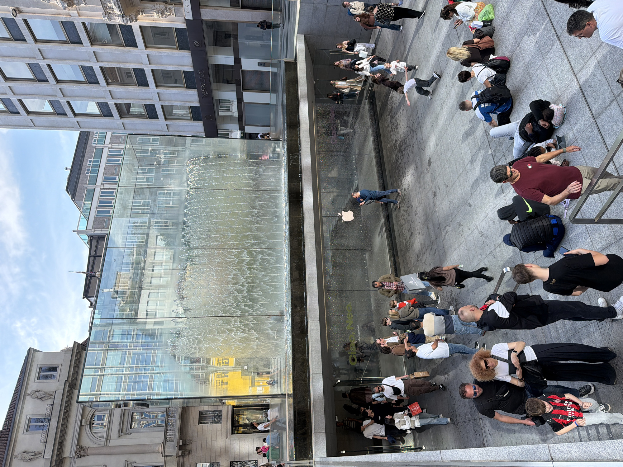 The image shows a crowd of people outside the Apple Store in Milan, Italy, likely drawn by a new product release. Many individuals are seated on the pavement, some possibly waiting in line. The scene is dominated by the store's modern glass facade and waterfall feature. The reflections on the glass reveal the surrounding architecture, including a building with "NESPRESSO" signage. The day appears sunny.