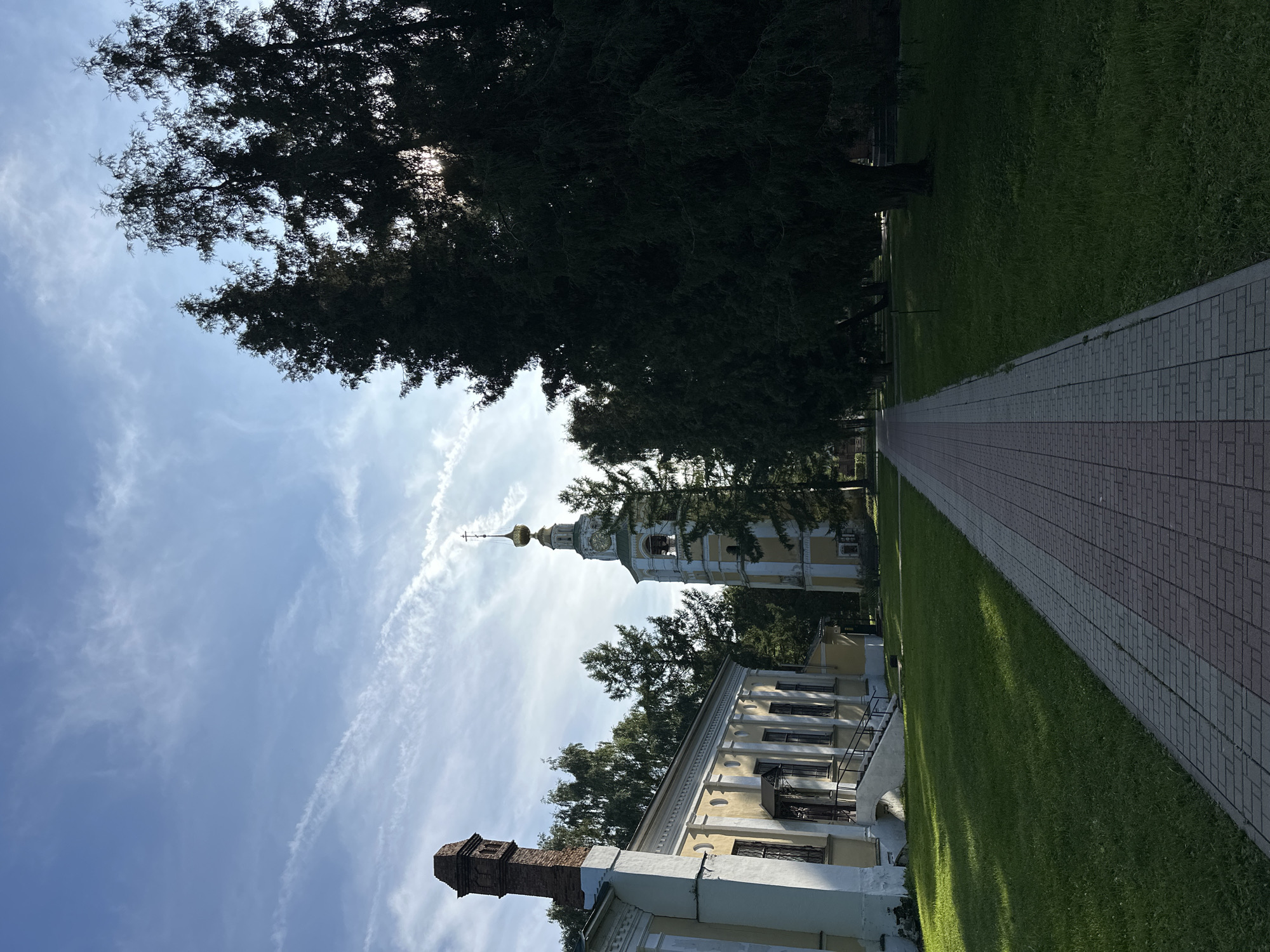 An exterior, eye-level, wide shot shows a paved walkway leading towards a yellow and white bell tower, topped with a gold dome and cross, situated in Selivanovo, Russia. The walkway is made of red and grey brick pavers and is flanked by a lush green lawn on the left and a dense line of dark green evergreen trees on the right. A long, low, yellow building with white trim and a red brick chimney is visible on the left side of the frame, casting long shadows across the lawn. The sky is a bright blue with scattered wispy white clouds. The sun is visible behind the trees on the right, creating a lens flare effect and dappling the ground with light. The scene appears serene and bathed in natural light.