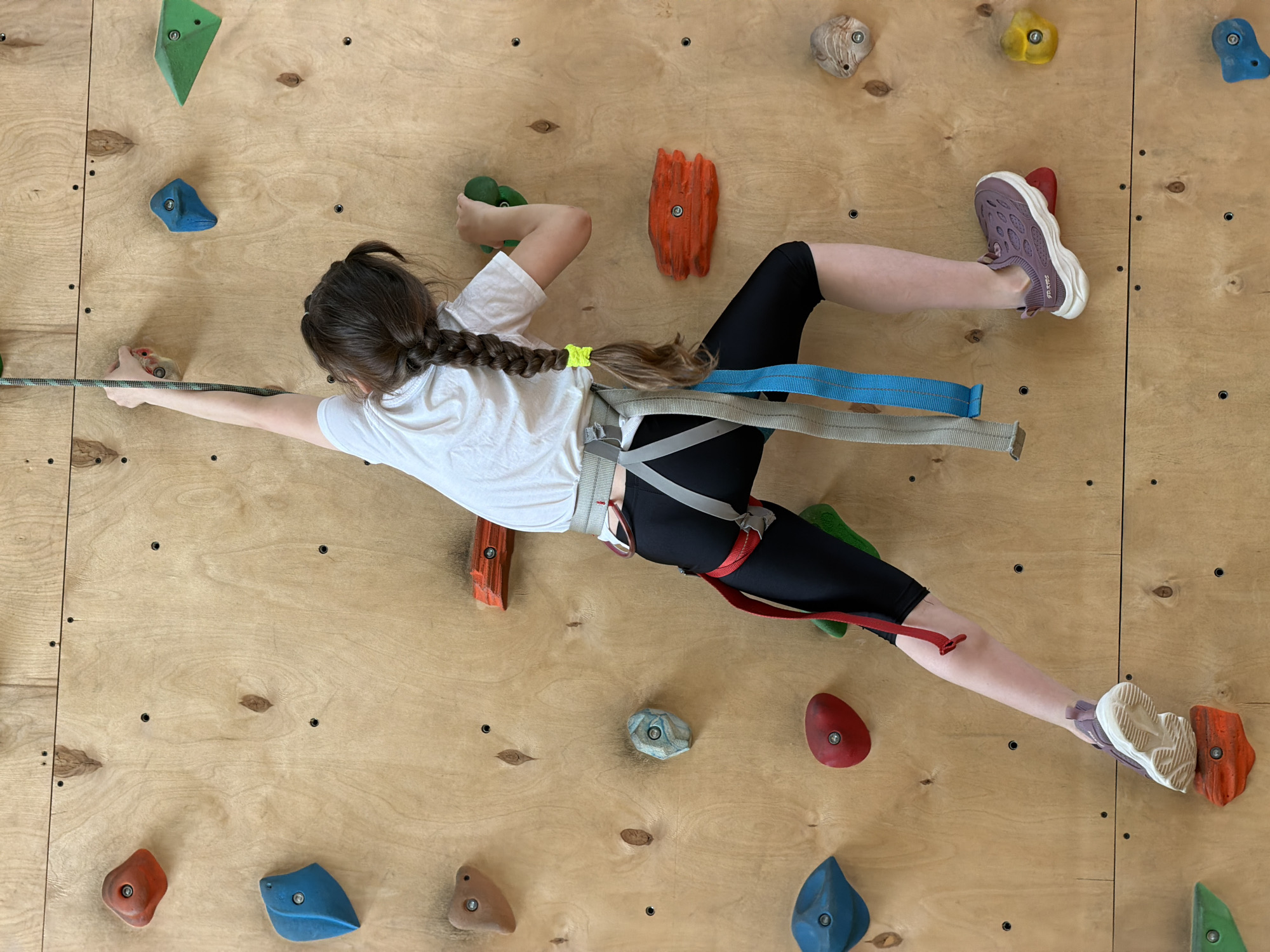 A young person is shown mid-climb on an indoor climbing wall. The individual is wearing a white t-shirt, black leggings, and athletic shoes, and is secured in a climbing harness. Their dark hair is styled in a braid with a yellow hair tie. The climber has one hand gripping a hold to the left and one foot resting on a hold to the right, seemingly in motion. The wall is made of wood with various colored climbing holds attached. The background is a plain wooden surface. The image does not provide sufficient information to confirm the location is in Izhevsk, Russia.
