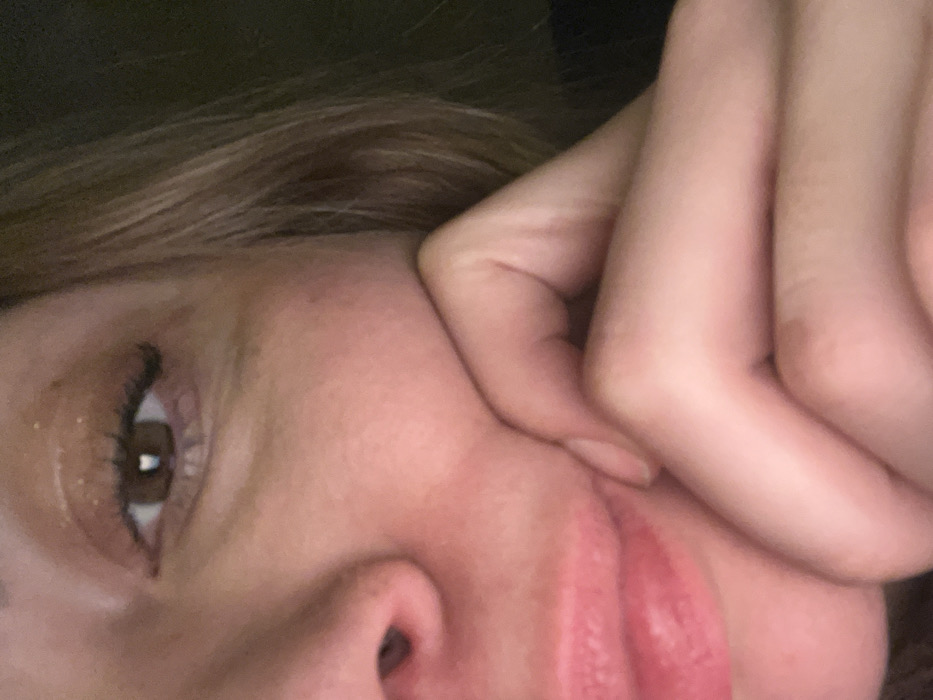 The image displays a close-up, low-light shot of a person's upper right face and right hand. The location cannot be determined from the visual information provided.
The subject is an individual with light-toned skin and light brown hair, strands of which are visible on the right side of the frame. The right eye is open, revealing a dark brown iris and a distinct white, heart-shaped reflection in the pupil. The eyelid features dark, elongated eyelashes, likely enhanced by mascara, and a light-colored, possibly shimmering, eyeshadow. Subtle lines are visible under the eye. The bridge and right nostril of the nose are partially visible on the left side of the frame. The lips, appearing full and naturally pink, are slightly parted in the lower-left quadrant of the image. The fingers of the right hand are positioned against the subject's lower right cheek and corner of the mouth, applying pressure and causing minor skin creases. The background is uniformly dark and out of focus, providing no environmental context. The scene is dimly lit, with a localized light source evident from the reflection in the eye.