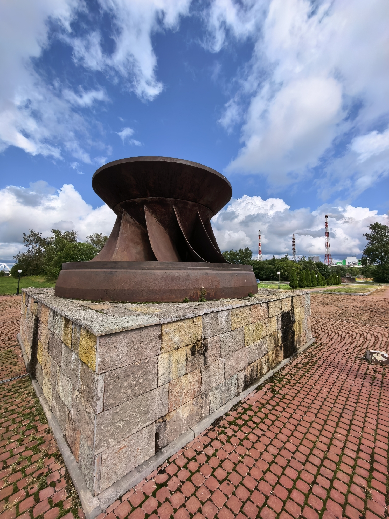 The image shows a large, rusted metal turbine component mounted on a stone pedestal. The turbine is dark brown and has a flared, bowl-like top with curved blades. The pedestal is constructed from irregularly shaped stones of various colors, including beige, yellow, and pink. The ground around the pedestal is paved with red brick pavers, with some patches of moss and grass growing between them.
In the background, there are several industrial chimneys with red and white stripes, emitting white smoke, indicating an active industrial site. Green trees and bushes form a line behind the chimneys, and the sky above is blue with scattered white clouds. The scene appears to be outdoors in a park-like setting, possibly commemorating industrial history.
This monument is located in the city of Volkhov, Russia, and is likely associated with the Volkhov Aluminum Plant or a similar industrial facility, given the presence of the chimneys. The turbine component itself suggests a connection to hydroelectric power or industrial machinery. The overall atmosphere is one of historical industrial presence set against a backdrop of a functioning industrial complex and a clear sky.