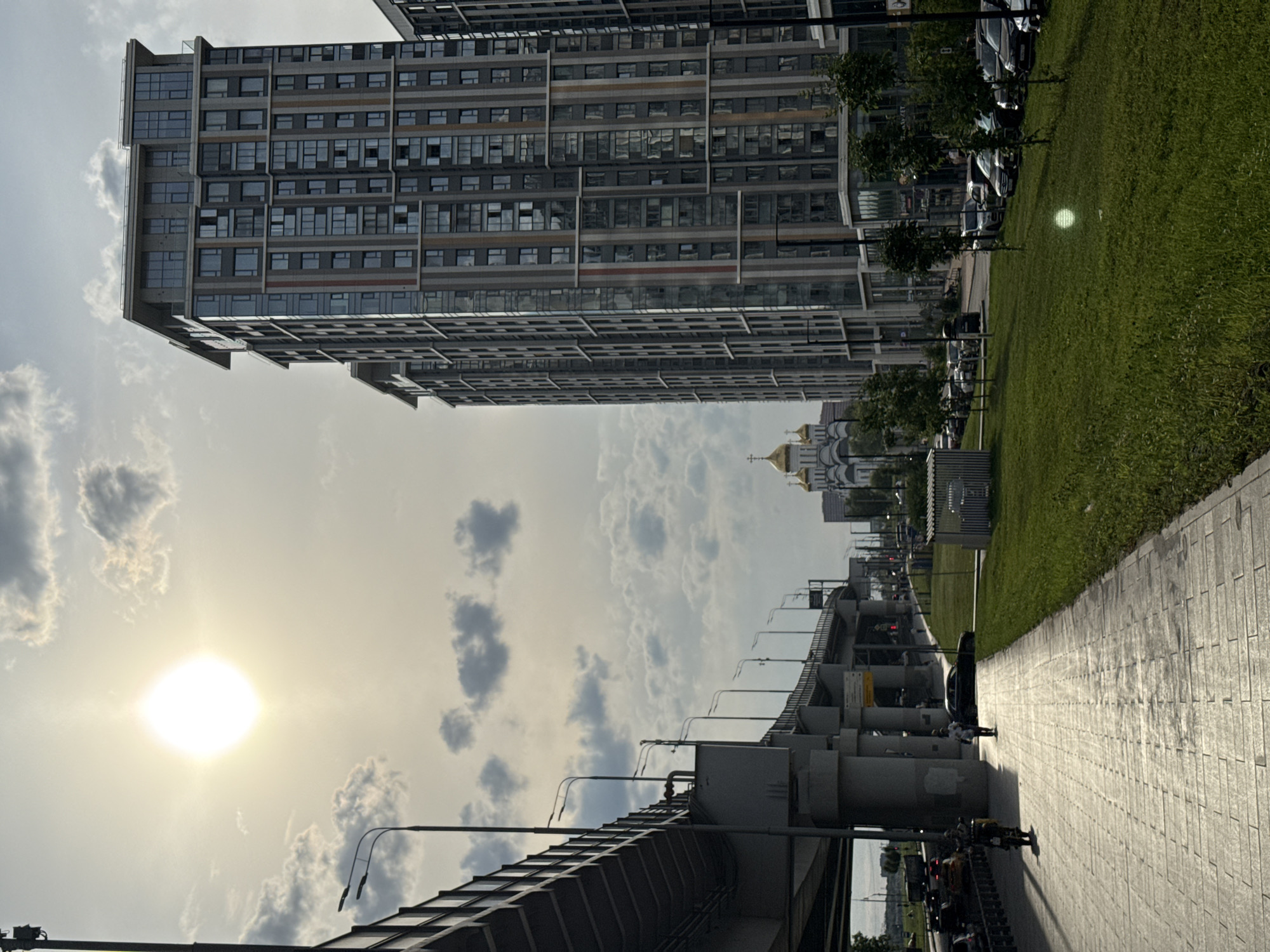 The image depicts a sun-drenched urban scene in Moscow, Russia. In the foreground, a sunlit, stone-paved walkway runs alongside a neatly manicured grassy area. Two individuals are walking on the walkway, one ahead of the other. To the left, a multi-level overpass with concrete pillars dominates the view, casting shadows on the walkway. Beyond the overpass, a street is visible with cars in motion and parked vehicles.
In the background, a modern, multi-story residential building with a grey facade and numerous windows stands prominently. To the left of this building, a classical-style church with golden domes is visible. The sky is partly cloudy with the sun shining brightly through the clouds, creating a lens flare effect in the center of the image. The overall atmosphere suggests a late afternoon or early evening scene in a developed urban area.