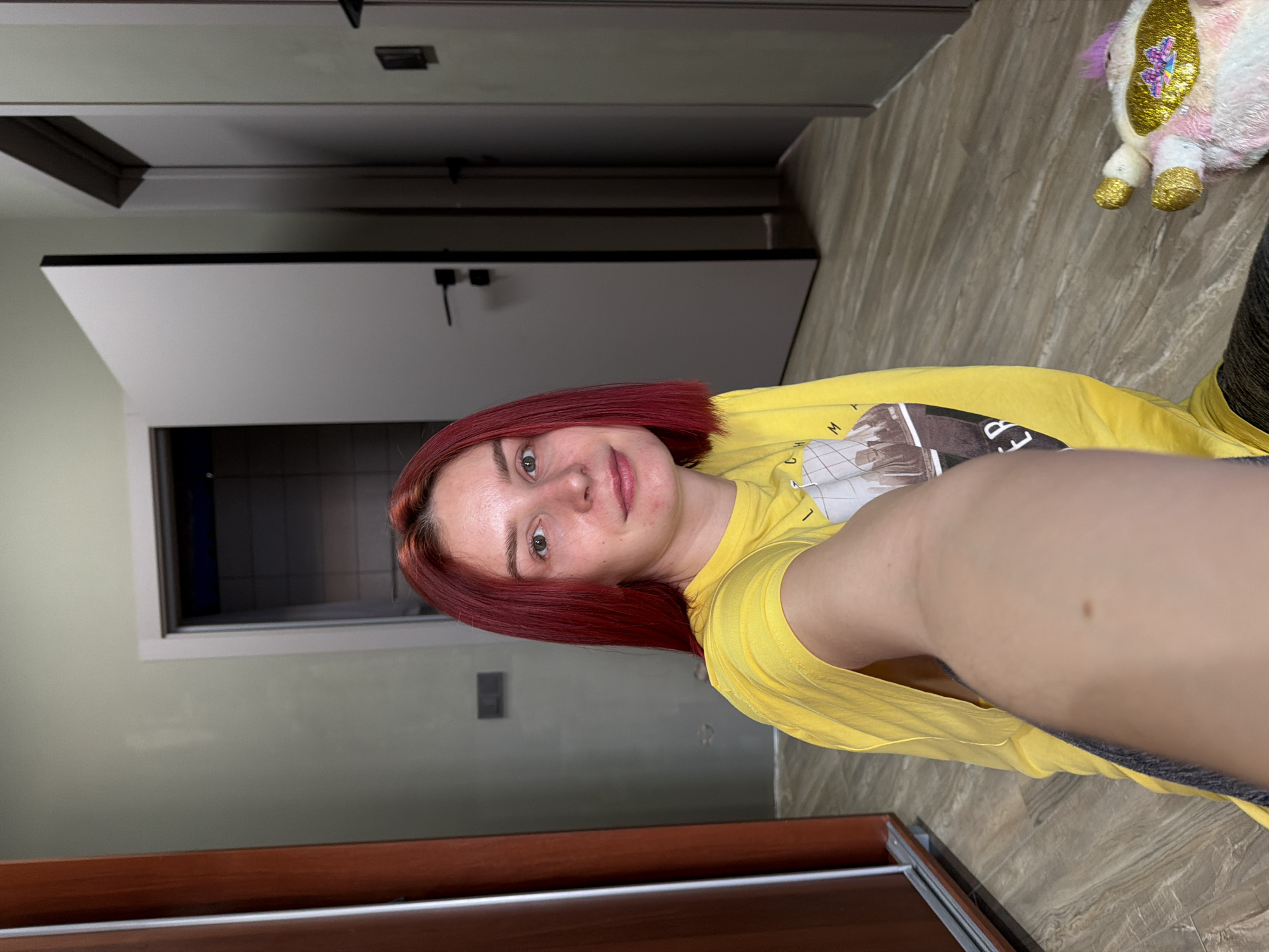 A young woman with short, reddish-brown hair is featured in a slightly rotated selfie taken indoors. She looks directly at the camera with a calm, subtle smile, displaying light-colored eyes and fair skin. She wears a yellow long-sleeved shirt with fragments of text, including "M," "CH," and "ER," visible alongside a graphic print. Her right arm, holding the camera, extends into the foreground.
The background depicts an interior room, likely a residential space. The floor is covered in light brown and grey patterned tiles or laminate. In the upper right corner, a colorful plush unicorn toy with gold glitter accents on its hooves and body is visible. Directly behind the woman, there is a white door with a black handle. Further to the left in the background, another dark-framed opening, possibly a doorway or window, leads to an adjacent room. The walls are painted a muted, light green or grey. A dark wooden piece of furniture, possibly a cabinet or wardrobe, is partially visible in the lower left foreground.