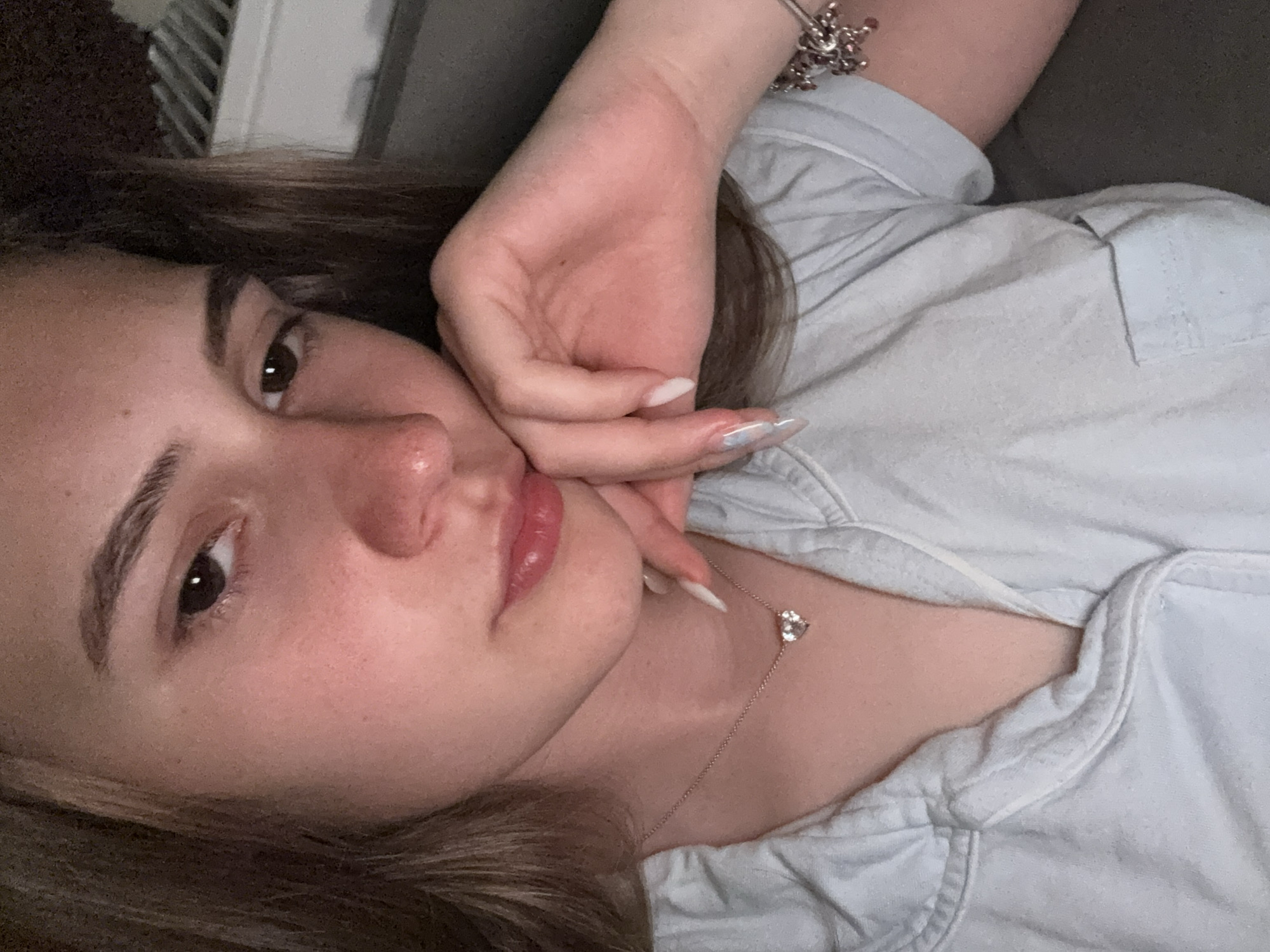 A young woman is lying on her side. Her head rests on her hand, which is placed near her face. She is wearing a light-colored, collared shirt and a necklace with a heart-shaped pendant. A bracelet is visible on her wrist. The background is out of focus, but part of a radiator can be seen. The location is unknown.