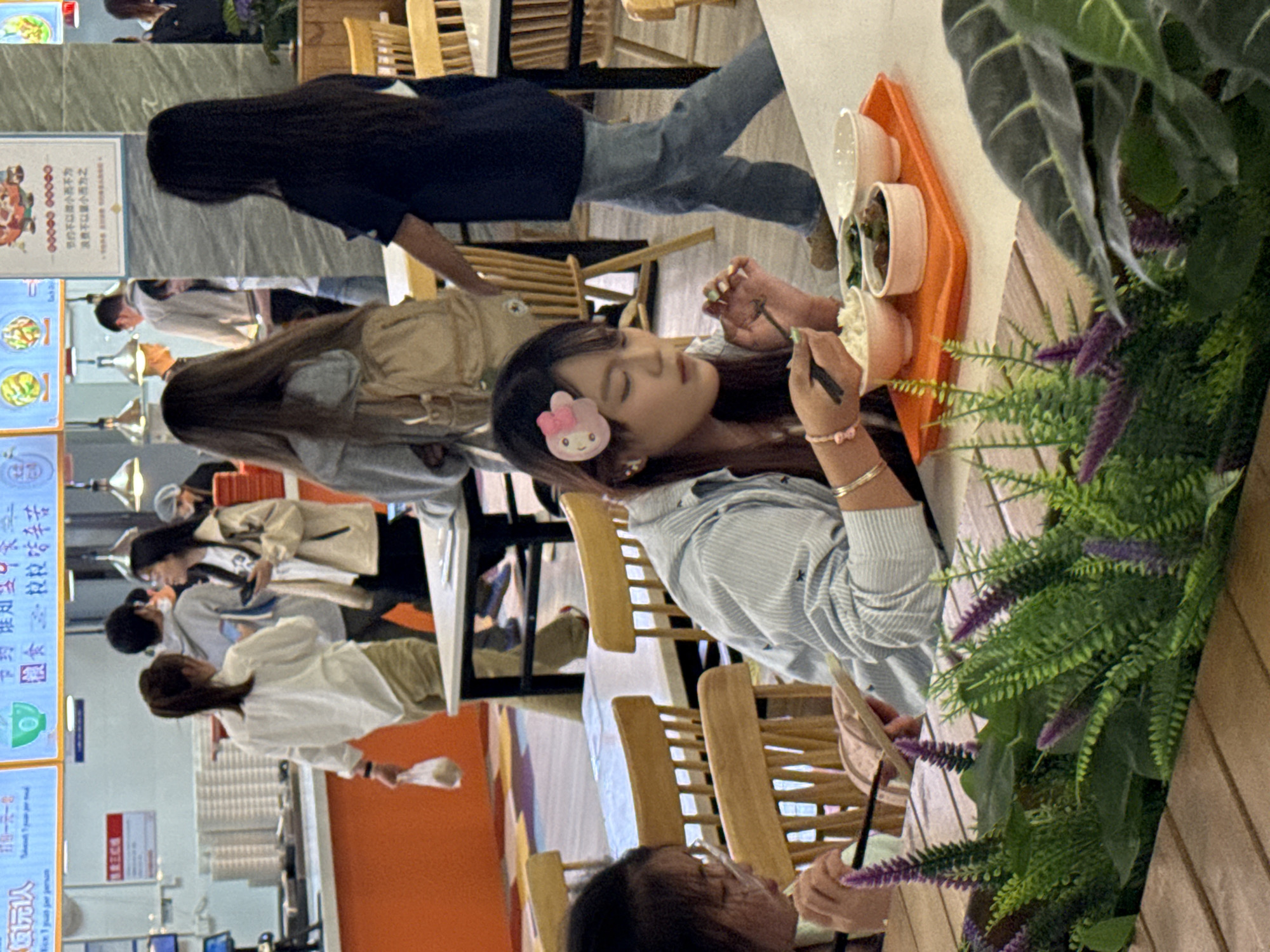 Here is a description of the image:
The image shows a scene at what appears to be a cafeteria, possibly in the city of Jiangjin, China. Several people are sitting at tables. One woman, positioned towards the right side of the frame, is using chopsticks to eat from a tray of food. She has a pink hair clip. Other individuals are seated around her. There are potted plants on a table next to her. In the background, a menu board is visible, featuring prices and descriptions of food items in Chinese characters. The setting seems to be a public dining area.