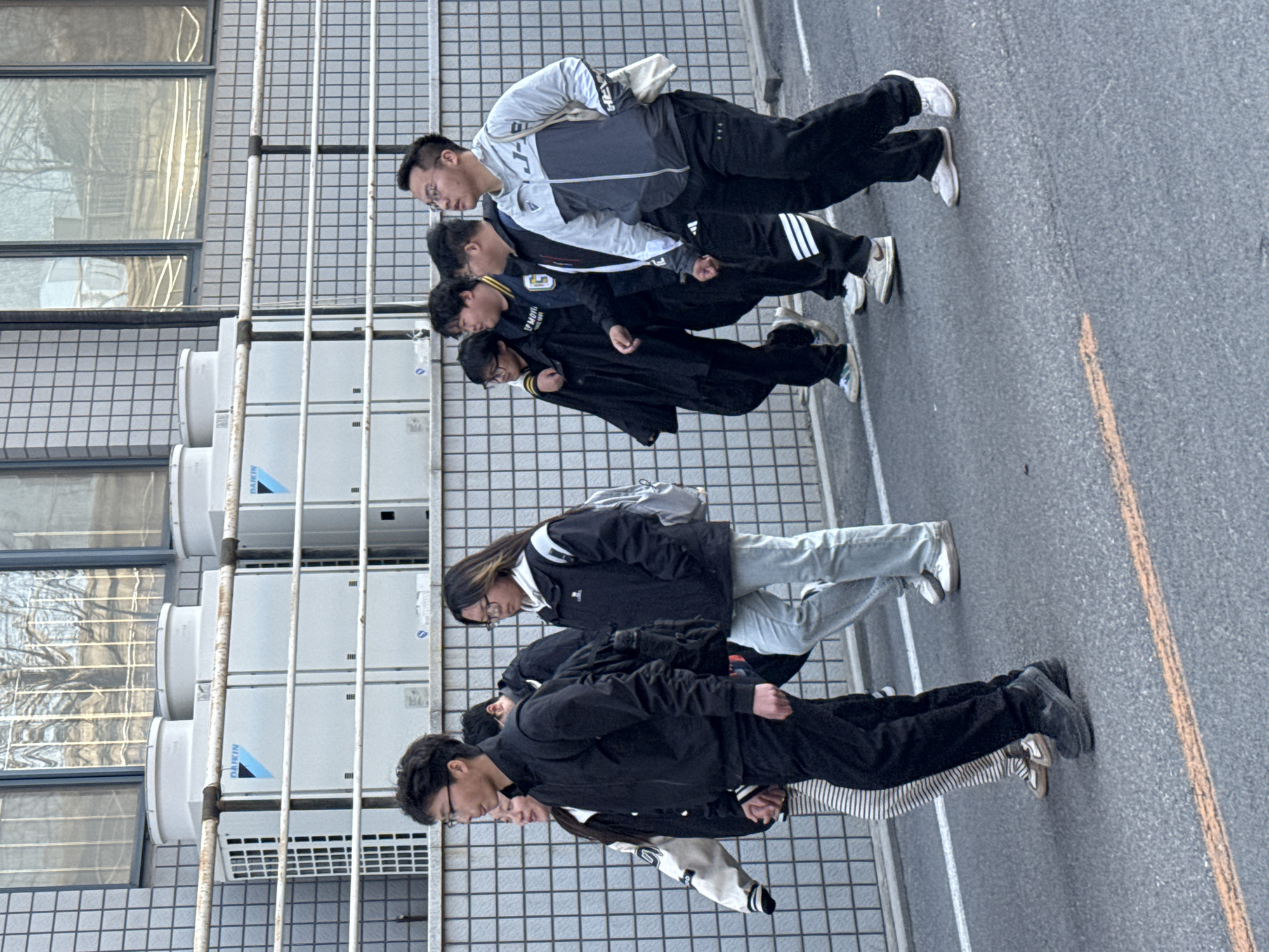 A group of approximately six people, likely students, are standing in a line along a street in Zhongtun, China. They are standing near the side of a building with multiple air conditioning units attached to its exterior wall. The street itself is paved and has yellow lines marking traffic lanes. The people are wearing a variety of clothing, including jackets, casual pants, and sneakers. Several appear to be wearing glasses. They are standing close together, possibly waiting for something or participating in an activity. The general environment suggests a public space, possibly near a school or educational institution. The image is taken at a slightly skewed angle.