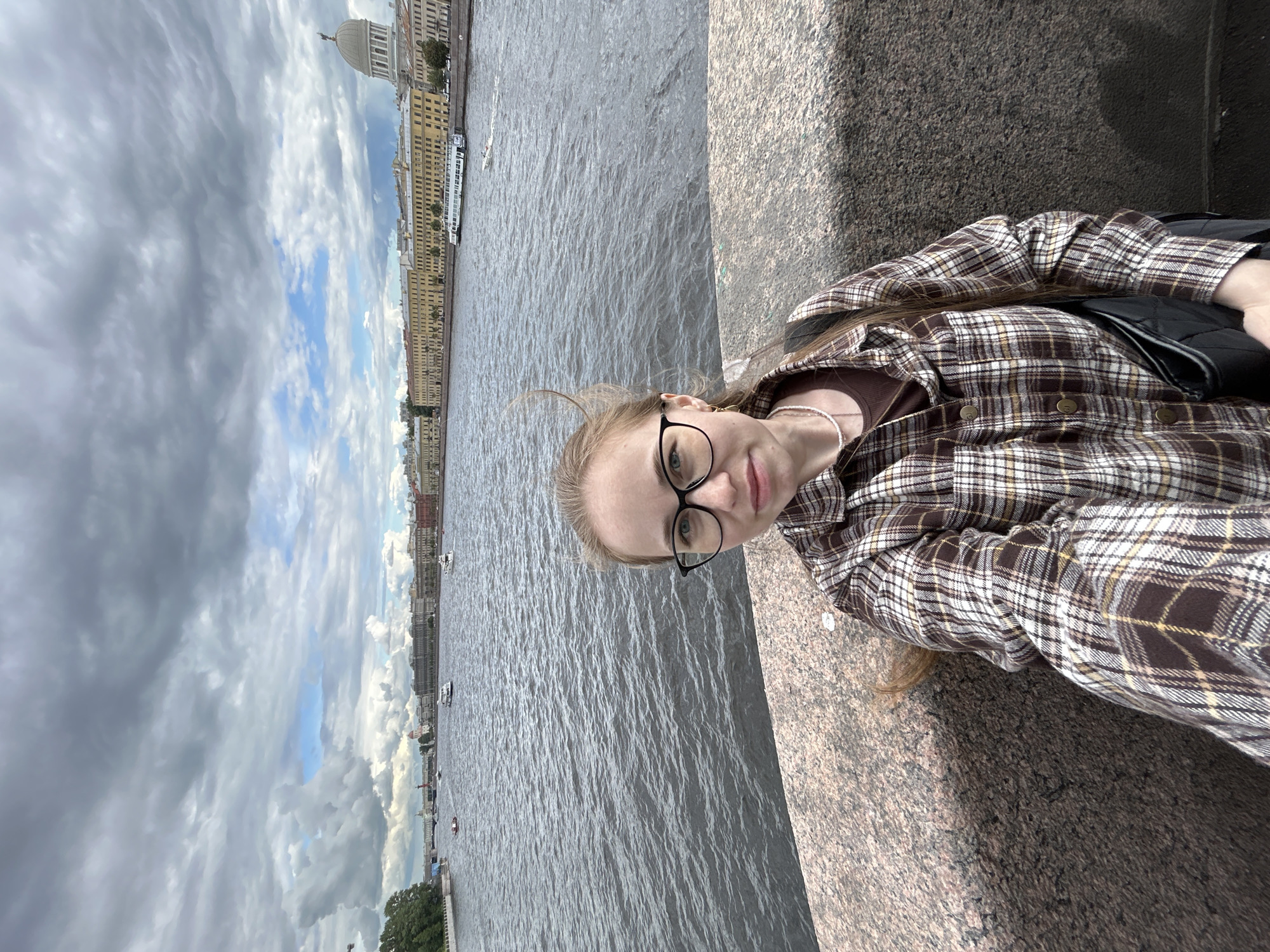 A young woman with blonde hair and glasses is taking a selfie outdoors, sitting on a granite ledge next to a body of water. She is wearing a brown and white plaid shirt over a brown top and a pearl necklace. The water appears to be a river or canal, with buildings and a dome structure visible on the opposite bank. The sky is cloudy with patches of blue sky. Several boats are on the water. The image is oriented vertically, with the woman on the right side of the frame. The scene appears to be during the day.