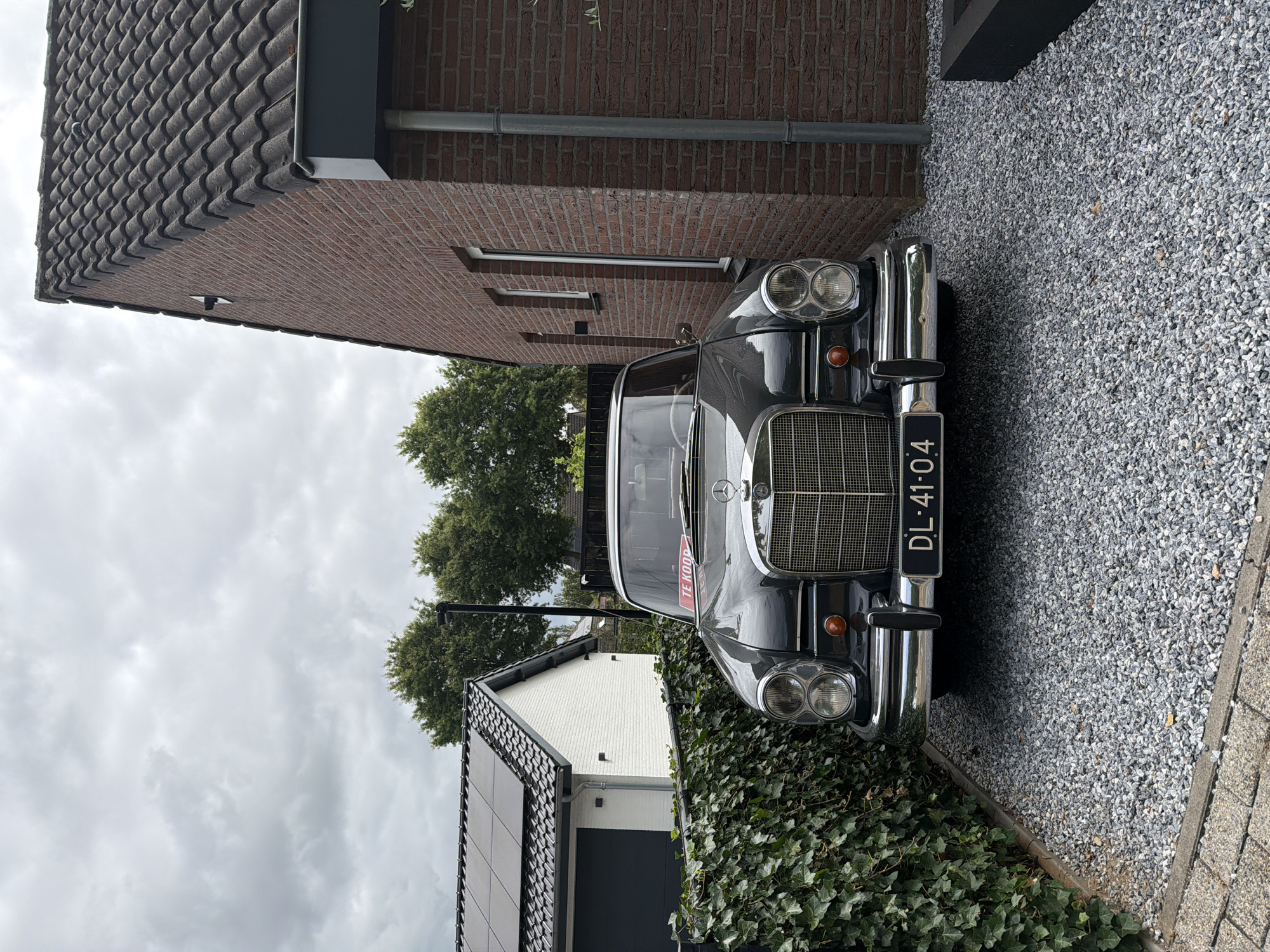 The image captures a vintage dark grey Mercedes-Benz sedan parked on a gravel driveway in front of a modern red-brick house. The car has a license plate reading "DL-41-04" and a red "TE KOOP" (for sale) sign is visible on the windshield. To the left of the car, a white building with a garage door and solar panels on its roof is partially visible, separated by a dense green ivy hedge. The scene is set under an overcast sky, with trees visible in the background. The location is identified as Melick, Netherlands.