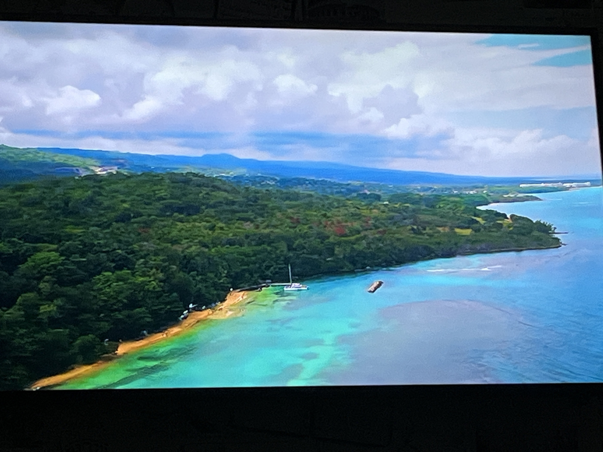 The image shows an aerial view of a coastline with lush green trees, a sandy beach, and clear turquoise water. In the water, there is a white sailboat and a brown rectangular object. The sky is filled with white and gray clouds. The geographical location is not Kharkiv, Ukraine, as the scenery depicted is characteristic of a tropical or subtropical coastal region.