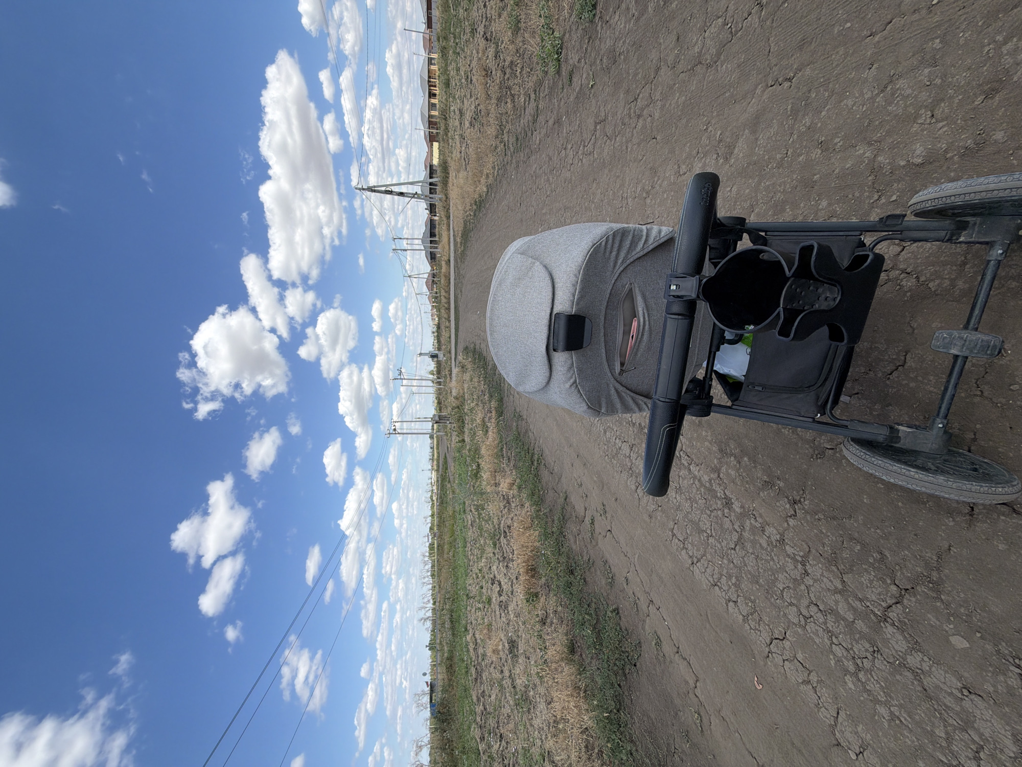 A first-person perspective shot shows a stroller parked on a dirt road. The stroller is grey and black, with a black cup holder attached to the handle. The road is cracked and dry, with sparse, dry grass growing on either side. In the background, a line of houses with tiled roofs stretches across the horizon. Utility poles with power lines are visible throughout the scene, suggesting a semi-rural or developing area. The sky is a vibrant blue, dotted with fluffy white clouds. The overall impression is of a sunny day in a somewhat undeveloped residential area. The location is identified as Krasnyi Krym, Russia.