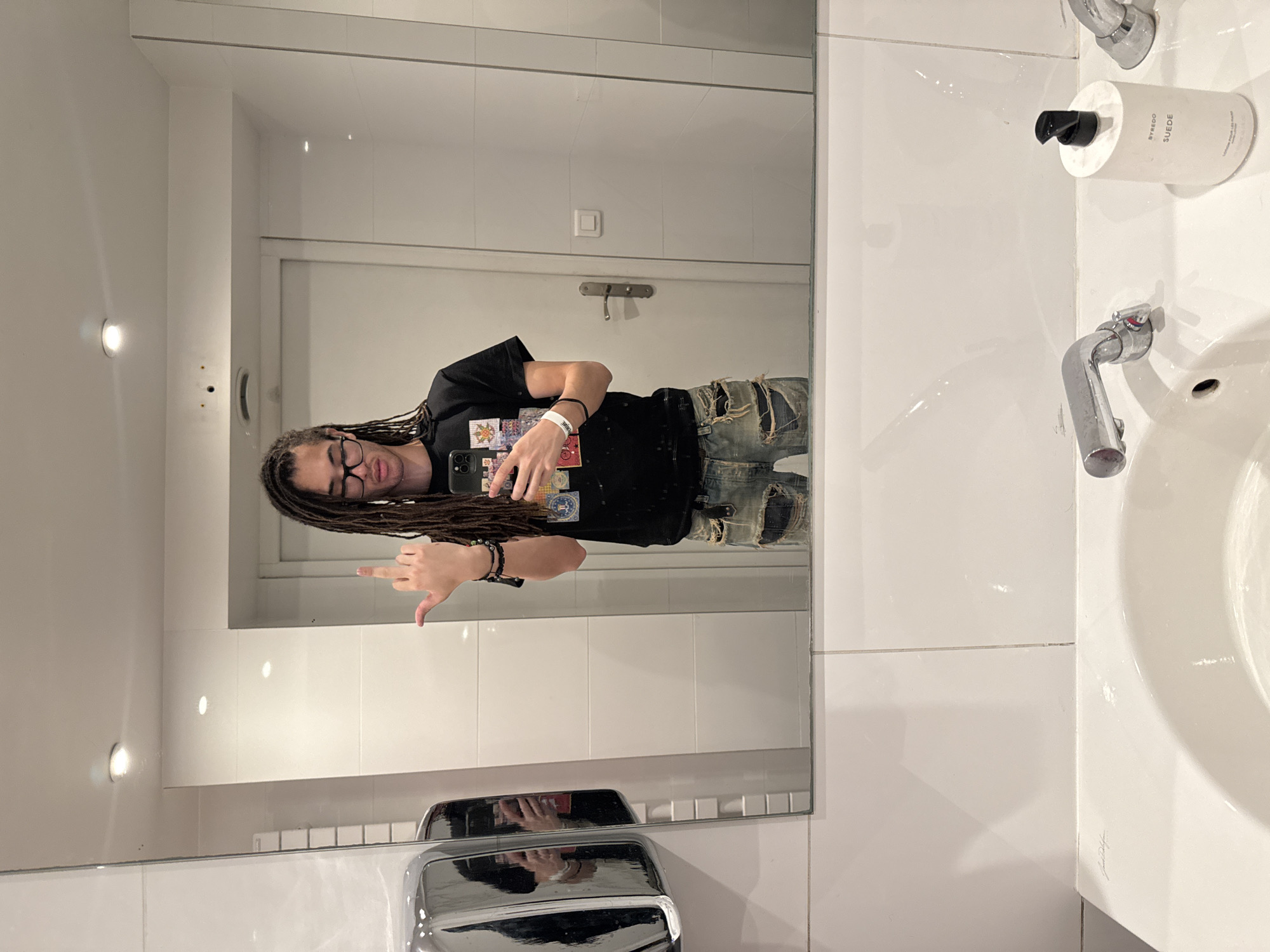 In a well-lit public restroom, a young person with dreadlocks and glasses stands in front of a mirror, holding a smartphone and making a rude gesture with their right hand. They are wearing a black t-shirt with a graphic print and distressed denim jeans. The restroom features white tiled walls, a silver hand dryer on the left, a sink with a chrome faucet, and a bottle of BYREDO SUEDE hand lotion. The scene is captured in a mirror reflection, suggesting the photographer is taking a selfie. The location is identified as Paris, France, based on the context provided.