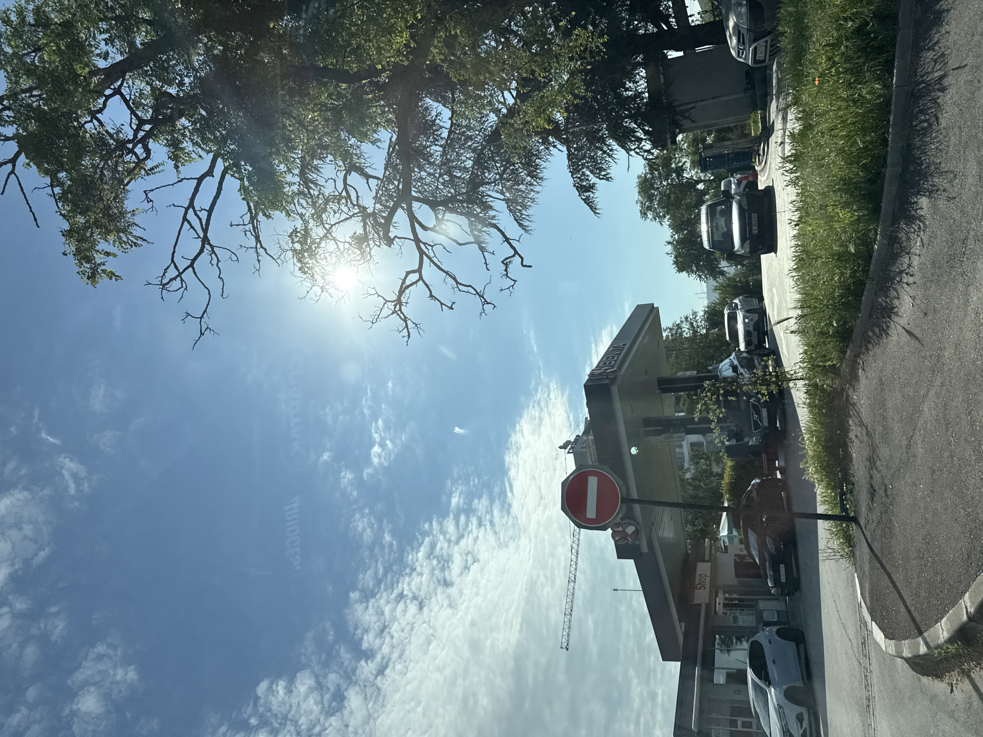The image depicts a gas station on a sunny day. A red and white "no entry" sign is prominently displayed in the foreground. Multiple cars are visible at the gas station, some appear to be refueling. A crane is visible in the background, suggesting construction in the vicinity. The sky is blue with scattered white clouds, and the sun is shining brightly. Lush green trees are present on the right side of the image, with some branches extending into the frame. The gas station has a "Shop" sign. The overall impression is a typical urban scene on a clear day.