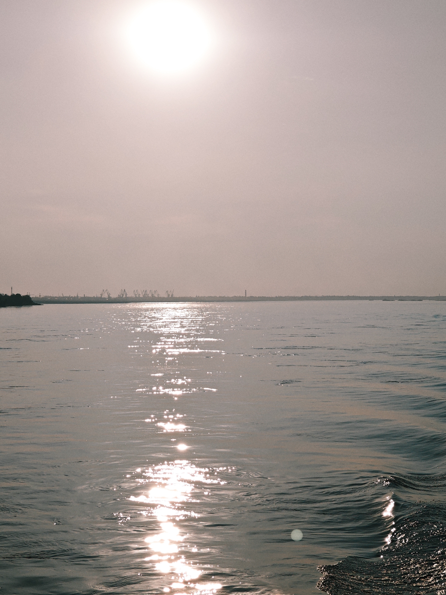 This is a wide-angle, vertical, eye-level shot of a body of water, likely the Angara River, in the city of Lesosibirsk, Russia. The sun is high in the hazy, light-pink sky, creating a bright, diffused light that casts a shimmering reflection on the water's surface. The water itself is calm with gentle ripples and waves, especially in the foreground where a wake is visible. The horizon line is low in the frame, with a distant shoreline visible. Silhouetted against the bright sky are several industrial cranes, suggesting the presence of a port or industrial area along the river. The overall atmosphere is serene and expansive.