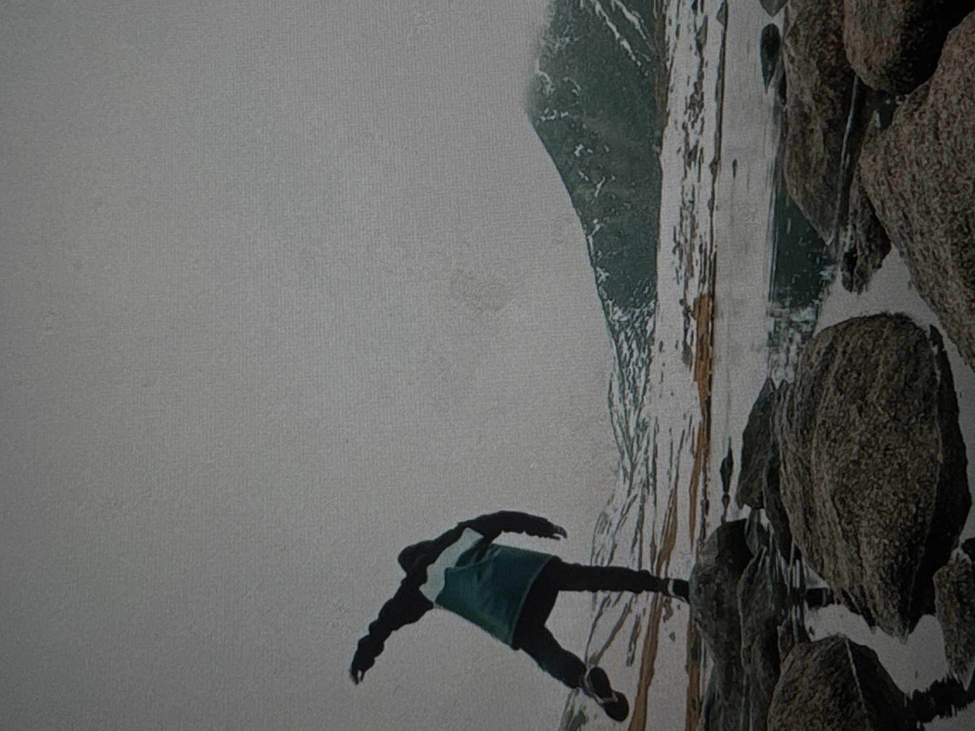 A person dressed in dark pants and a green and white jacket is captured mid-stride, with their left leg extended behind them and their right leg bent. Their left arm is raised and extended outwards, while their right arm is bent at the elbow. The individual is standing on a cluster of large, dark rocks that are partially submerged in shallow water. The surrounding landscape is bleak and appears to be wintry, with snow-covered ground and hills in the background. A large, dark mountain with patches of snow dominates the right side of the frame. The sky is overcast and a uniform gray. The scene is likely outdoors, in a natural environment. There is no information to suggest this image is related to the city of Irkutsk, Russia.