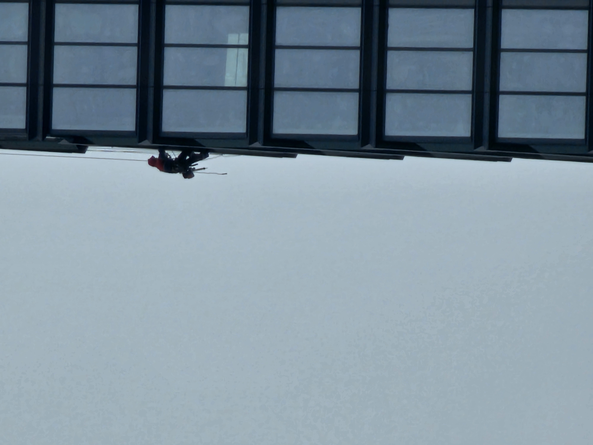 An image depicts a person suspended horizontally against a large, uniform light blue-grey background, which makes up the majority of the frame. In the upper portion of the image, a section of a modern building facade is visible, featuring multiple rectangular windows with dark frames.
The subject is an individual, likely a worker, wearing a red hooded jacket and dark trousers. They are equipped with harnesses and ropes, which are visible extending from their body and up towards the building. Their posture suggests they are either rappelling or performing rope access work on the exterior of the building. Given the horizontal orientation of the windows and the person, the image appears to be rotated 90 degrees clockwise from its likely original, upright perspective where the person would be vertically descending the building.
The light is soft and diffused, indicating daytime conditions. There are no discernible features in the background other than the uniform light tone, suggesting a clear sky or a distant, featureless surface. No text is visible in the image. The described location context is Moscow, Russia.