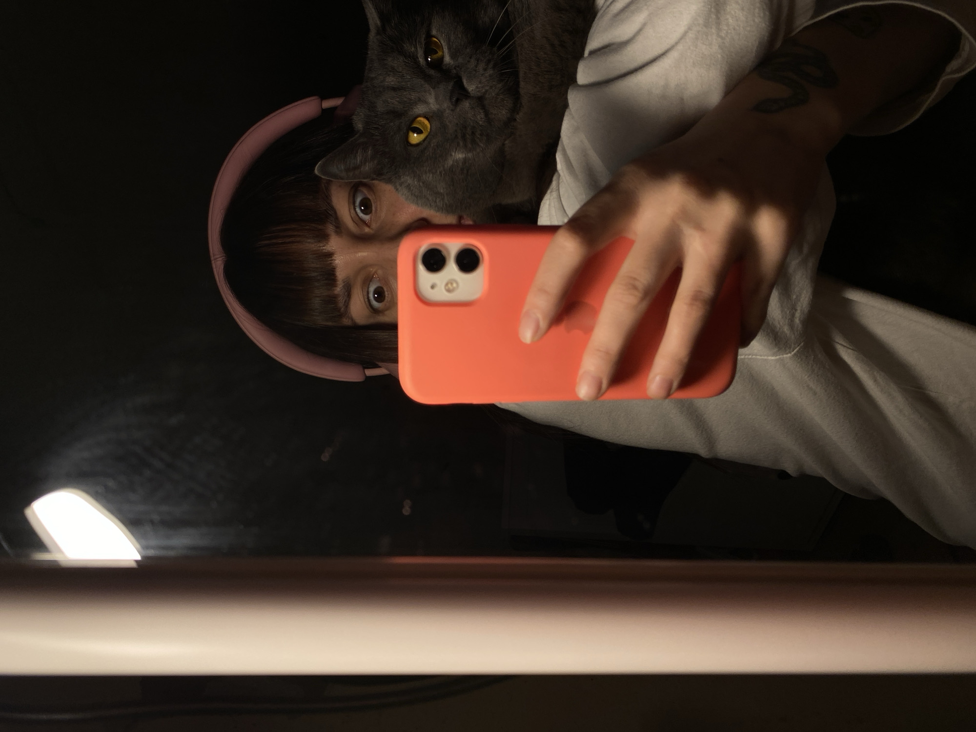 A low-angle, close-up selfie featuring a person and a gray cat. The person, wearing pink headphones, has short, dark hair with bangs and is holding an orange iPhone. The cat, with yellow eyes, is positioned near the person's face. The person is wearing a light gray shirt and has a tattoo on their left forearm. The background is dark, with a partially visible white table and a lamp illuminating the lower left corner. The location is not discernible.