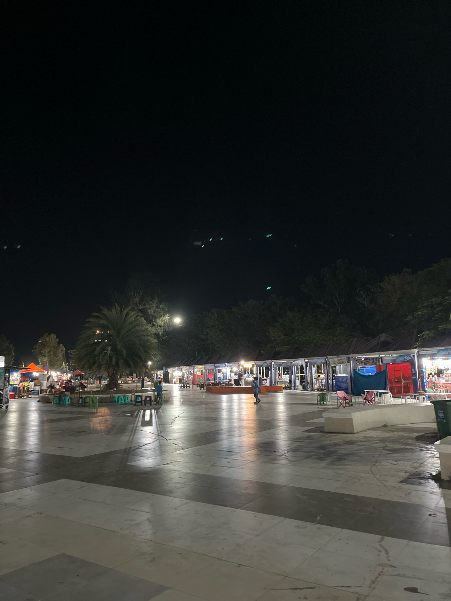 The image depicts a night scene at the Kendari City waterfront area in Kendari, Indonesia, likely near a night market or food court. In the foreground, a large, polished concrete plaza with patterned grey sections reflects the ambient light. Several small, colorful plastic stools are scattered throughout the plaza, some clustered around a central palm tree with a circular concrete planter.
In the midground, a row of open-air stalls with dark, angled roofs lines a paved area. These stalls are illuminated, showcasing various goods and food items, with some tables and chairs visible in front of them. A few individuals are present, appearing to be customers or vendors. One person in a blue shirt is walking across the plaza from right to left. Another person, a child, is seated on a stool. The background is dominated by trees and the dark night sky. A bright light, possibly a full moon or a strong artificial light source, is visible in the upper right portion of the image, casting shadows on the plaza. There are also faint, streak-like light artifacts in the sky.
