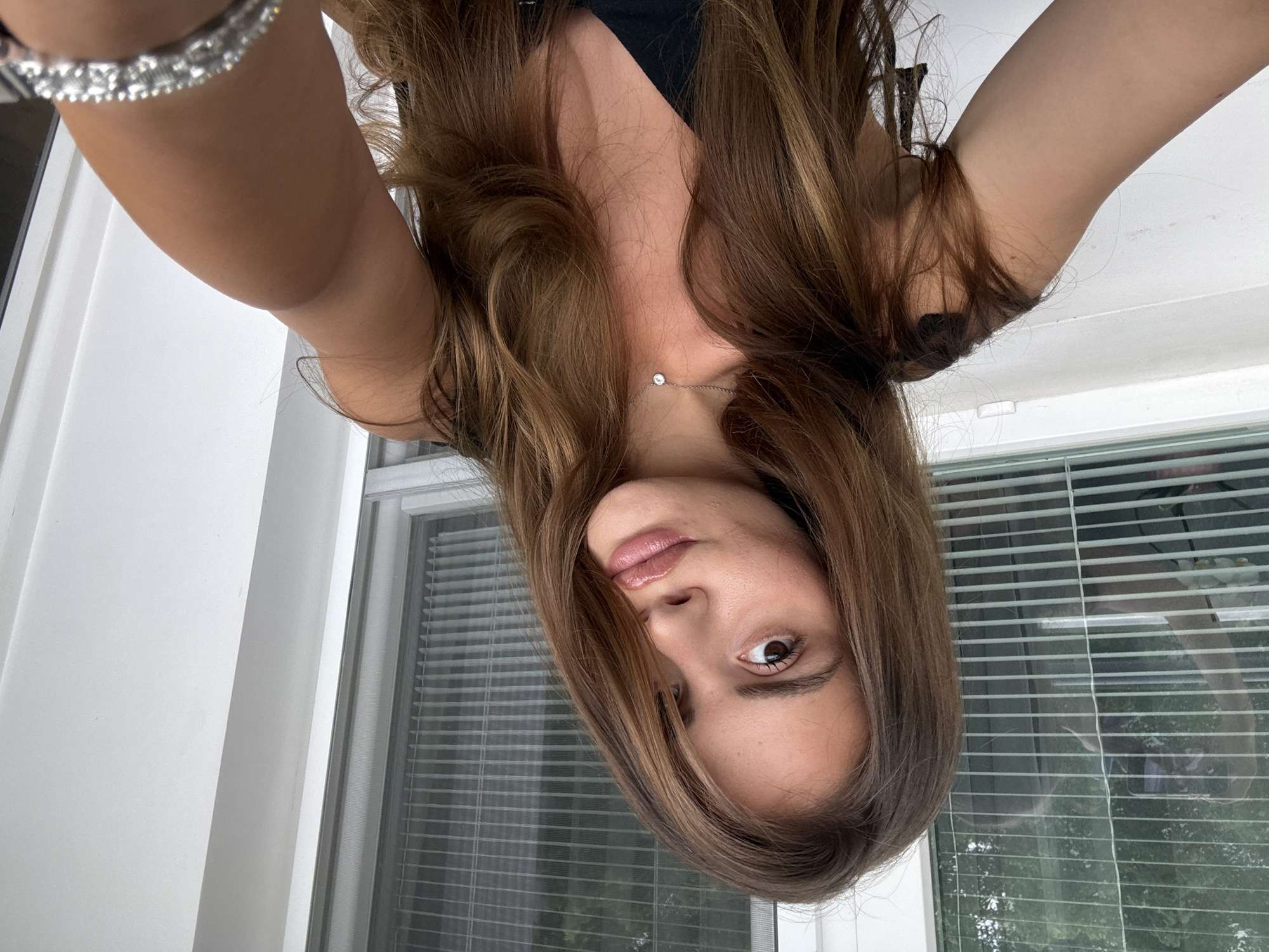 A young woman with long, brown hair takes a selfie. She is wearing a black top and a delicate silver necklace. Her right arm is raised, holding what appears to be a phone, though it is not fully visible. She has makeup on, including lip gloss and eyeliner. In the background, there is a window with horizontal blinds and a white wall. The lighting suggests it is daytime. The image is a personal photograph, likely taken indoors, potentially on a balcony or near a window. It is not possible to determine the location within the city of Plovdiv, Bulgaria, from the visual information provided in the image.