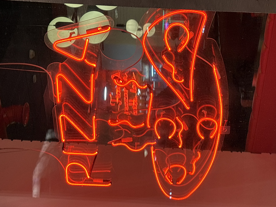 A bright red neon sign is displayed behind a glass storefront window, likely a pizza restaurant, located at Popova Street, 16, in Perm, Russia. The sign prominently features the uppercase word "PIZZA" at the top. Below this, a smaller, cursive neon inscription reads "From our ovens with love." Underneath the text, the sign depicts a stylized graphic of a whole pizza on the left, with an individual slice of pizza to its right, all rendered in red neon tubing. Reflections on the glass surface obscure some background details but reveal elements of the interior, including a visible red pipe, possibly part of a fire suppression system, and indistinct circular light sources or reflections from within the establishment. The lower portion of the image shows a red surface reflected in the window, suggesting interior furnishings. No individuals are visible. The scene is illuminated primarily by the vibrant red glow of the neon sign against a darker background.