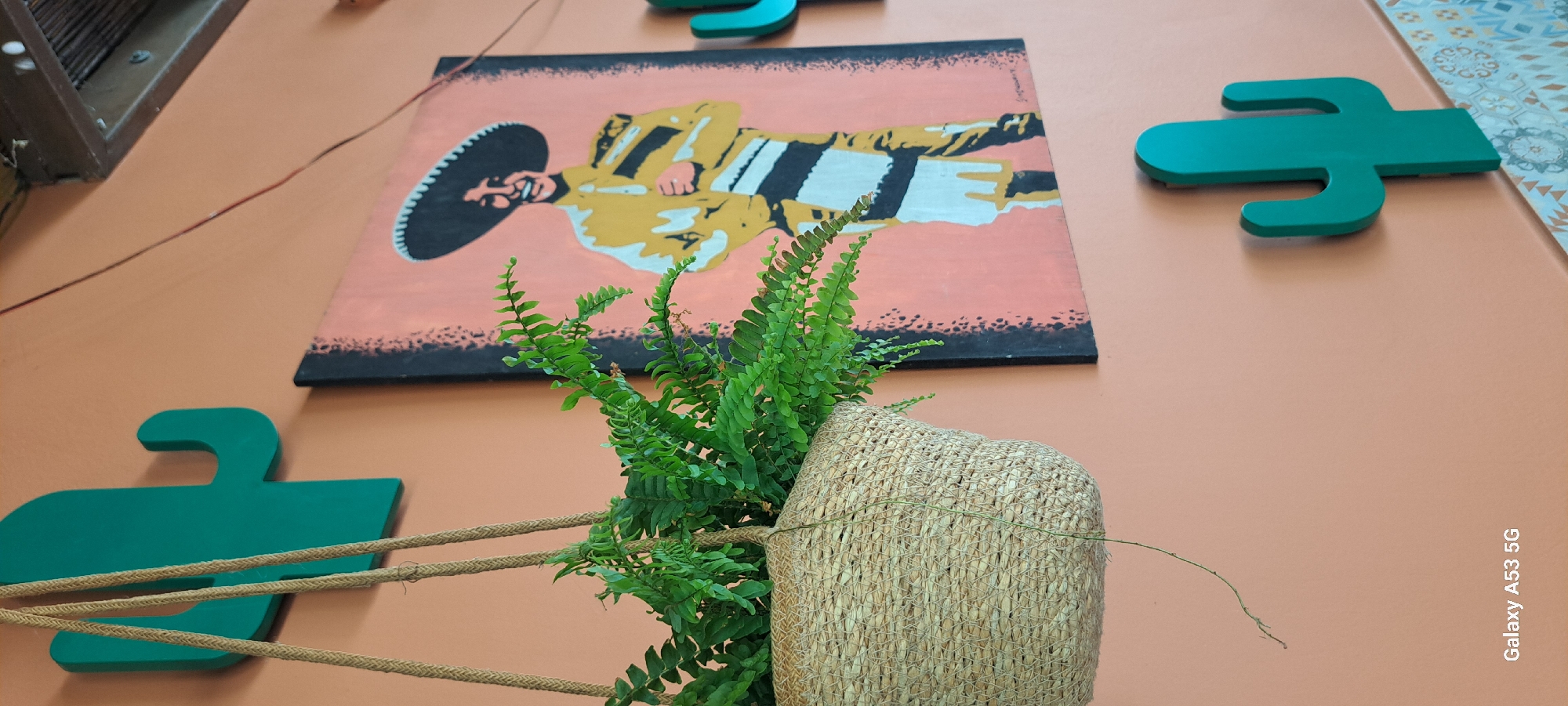 The image depicts a wall decorated with a painting of a mariachi musician, two green cactus-shaped cutouts, and a hanging fern in a woven basket. The painting features a smiling man in a wide-brimmed sombrero and a yellow outfit with black and white striped accents, set against a coral pink background. The cactus cutouts are positioned on the left and right sides of the painting, with the one on the left partially obscured by the hanging plant. The fern is lush and green, cascading from its basket, which is suspended by rope. The wall itself is painted a warm, peachy-orange color. The lower right corner of the image reveals a patterned tiled border. A thin wire runs diagonally across the upper right portion of the frame. The bottom left corner displays "Galaxy A53 5G", indicating the device used to capture the photo. The overall scene suggests a vibrant and possibly themed interior space, though its specific location in Kos, Greece, cannot be determined from the visual information provided.