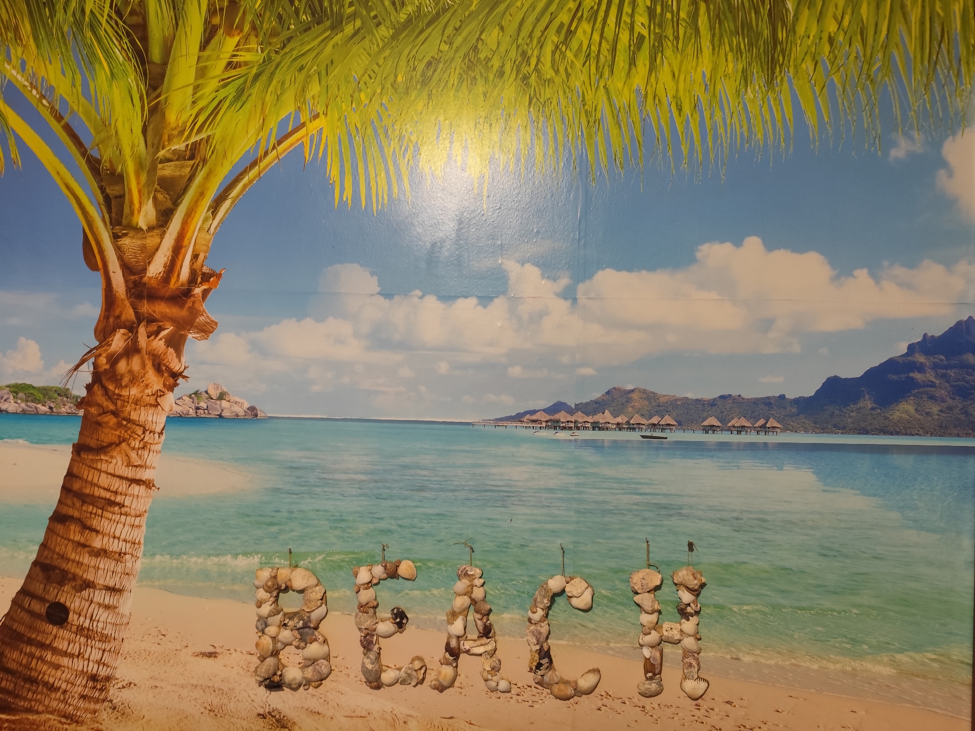 The image is a wide shot of a tropical beach scene. In the foreground, on the sandy shore, are the letters "BEACH" spelled out with seashells. The letter "B" is to the left, followed by "E", "A", "C", and "H". To the left of the letters is the trunk and base of a palm tree with its fronds extending across the top of the image. The middle ground features clear turquoise water with gentle waves lapping at the shore. In the distance, over the water, are several overwater bungalows with thatched roofs, forming a resort. Beyond the bungalows, a calm body of water stretches to the horizon, where distant mountains rise. The sky is a clear blue with scattered white clouds. The image depicts a serene and idyllic beach setting, likely a representation of a resort or vacation destination.