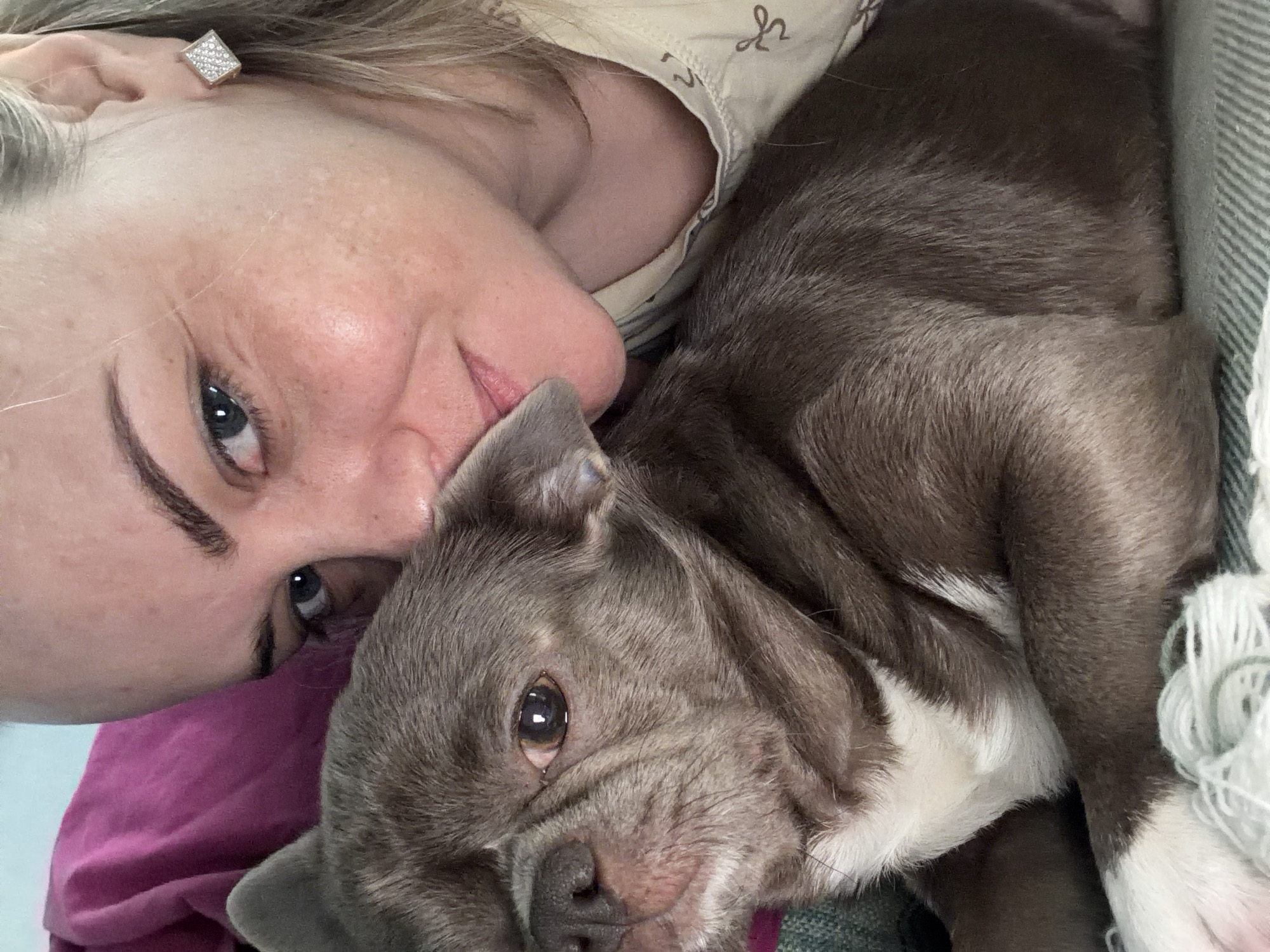 A woman with fair skin and blonde hair is lying down, closely embracing a brindle-colored dog with white markings on its chest and paws. The woman is wearing a light-colored top with a pattern, and a square earring is visible in her left ear. The dog's head is resting near her cheek. The background includes glimpses of a pink garment and a textured, possibly knitted or woven, surface. There is no evidence to suggest the image was taken in Votkinsk, Russia, or anywhere else specifically.