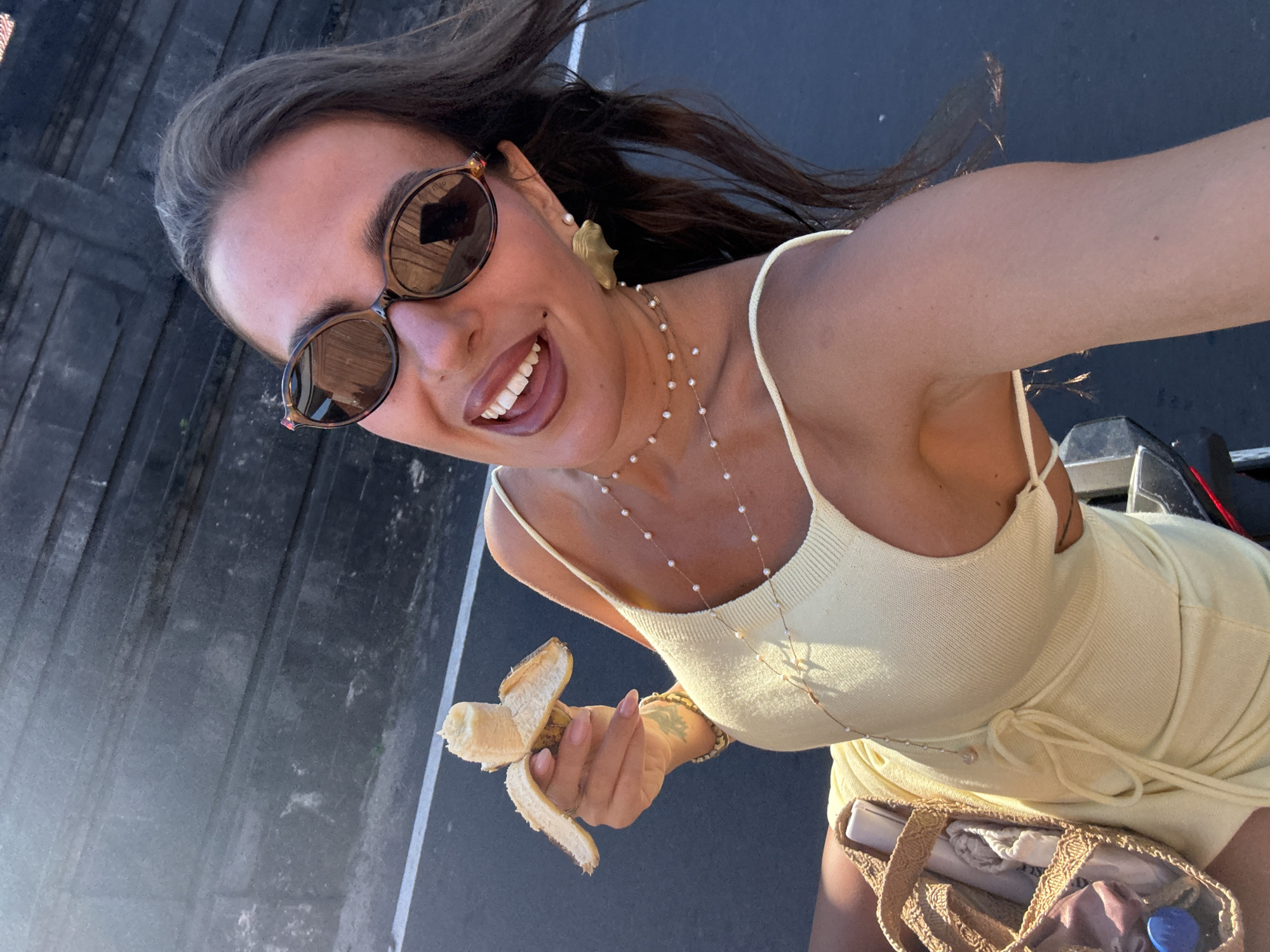 The image is a selfie of a woman with brown hair, wearing sunglasses and a light yellow dress, holding a partially peeled banana. She's smiling broadly. She also wears a pearl necklace and gold shell earrings. The background suggests an outdoor urban environment with a concrete or asphalt surface. Based on the prompt, the location is assumed to be Kuta Selatan, Indonesia, though there are no specific landmarks visible in the frame to confirm this.