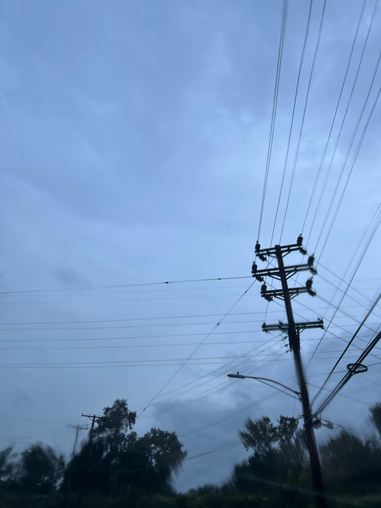 A utility pole stands against a cloudy, overcast sky, its dark silhouette sharply defined by the faint ambient light. Numerous power lines stretch across the frame, some taut and parallel, others angled and intersecting, creating a complex network against the muted blue-gray backdrop. The pole itself is a dark, vertical structure with horizontal crossarms, each supporting insulators and the wires. The lower portion of the image shows a blurred line of dark green trees and foliage, suggesting the pole is situated along a street or road. There are no people visible in the image. The overall impression is one of a typical urban or suburban landscape under a somber sky. The location is Scott Township, United States of America.