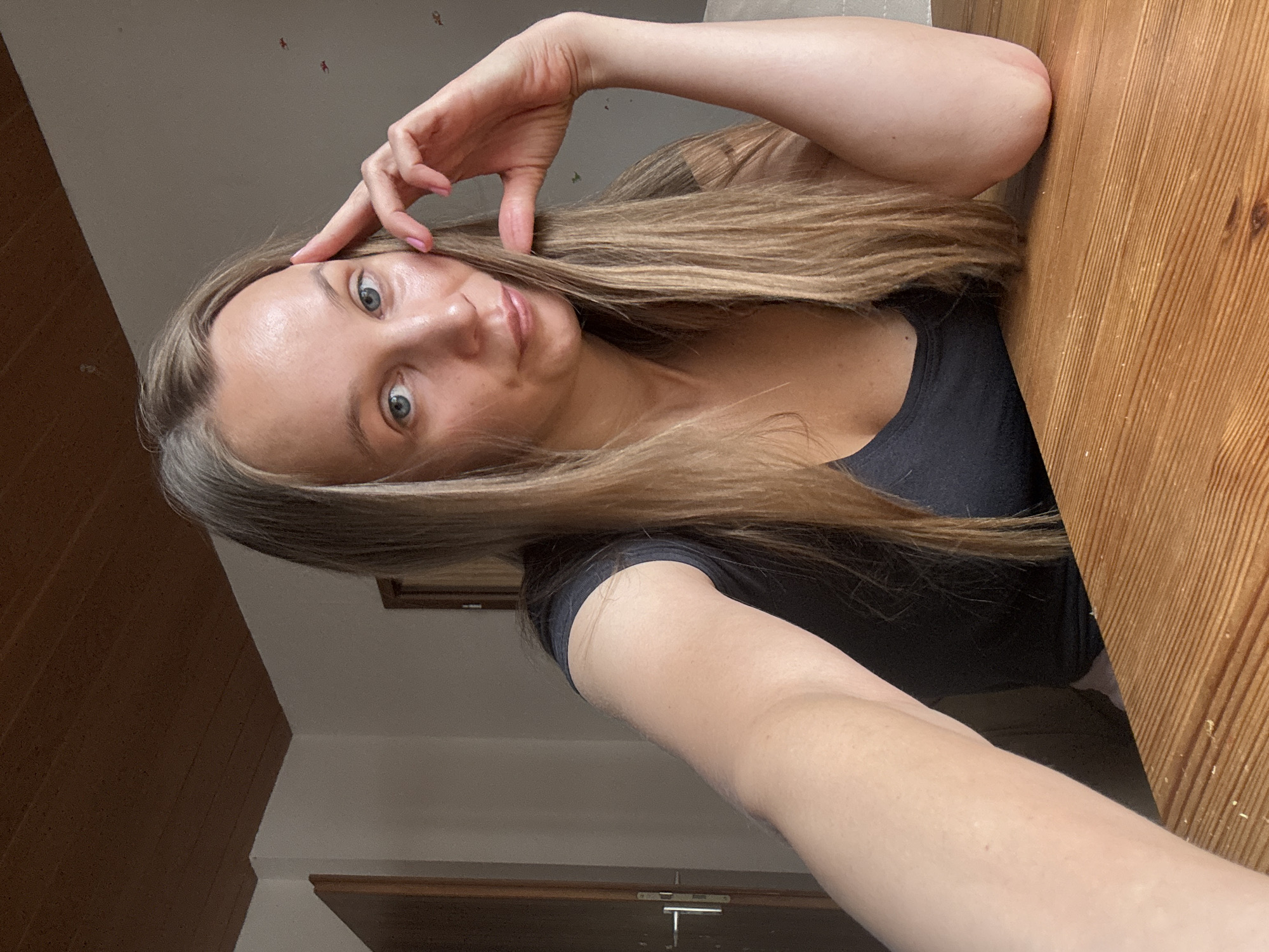 A woman with long blonde hair and blue eyes is sitting at a wooden table, looking towards the camera. She is resting her right hand on her forehead and her left arm is bent at the elbow, resting on the table. She is wearing a dark t-shirt. The background shows a room with a door and a ceiling made of wooden panels. The location is not identifiable as Überherrn, Germany.