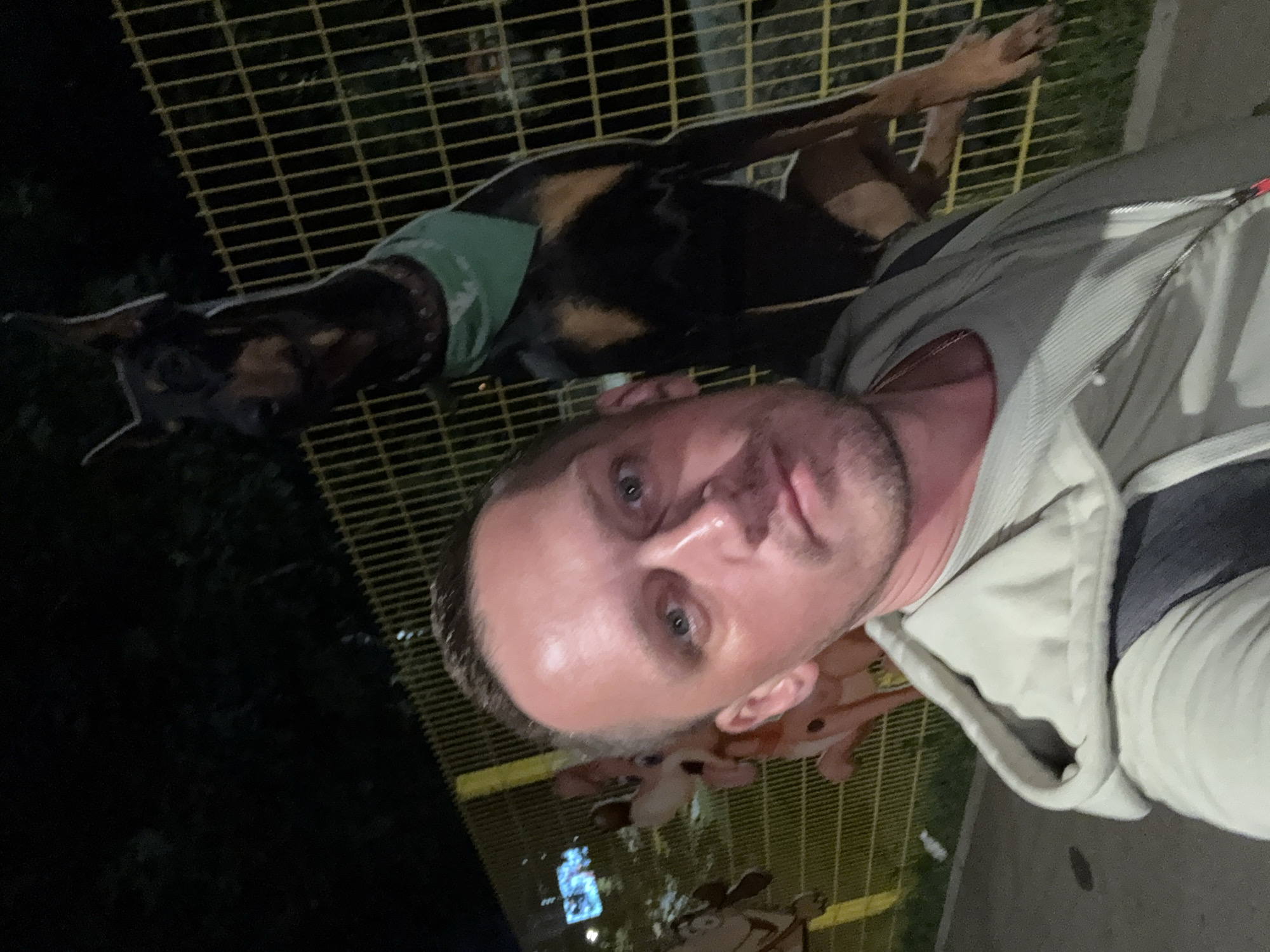 In an outdoor nighttime scene, a man with light skin and short brown hair is taking a selfie. He is wearing a light-colored hooded jacket over a grey t-shirt and a necklace. To his right, a Doberman Pinscher with black and tan fur is visible behind a yellow metal fence. The dog is wearing a green bandana around its neck and appears to be standing on its hind legs, with its front paws on top of the fence. In the background, blurry cartoon-like figures are attached to the fence, and dark foliage is visible. The ground in the foreground is paved.