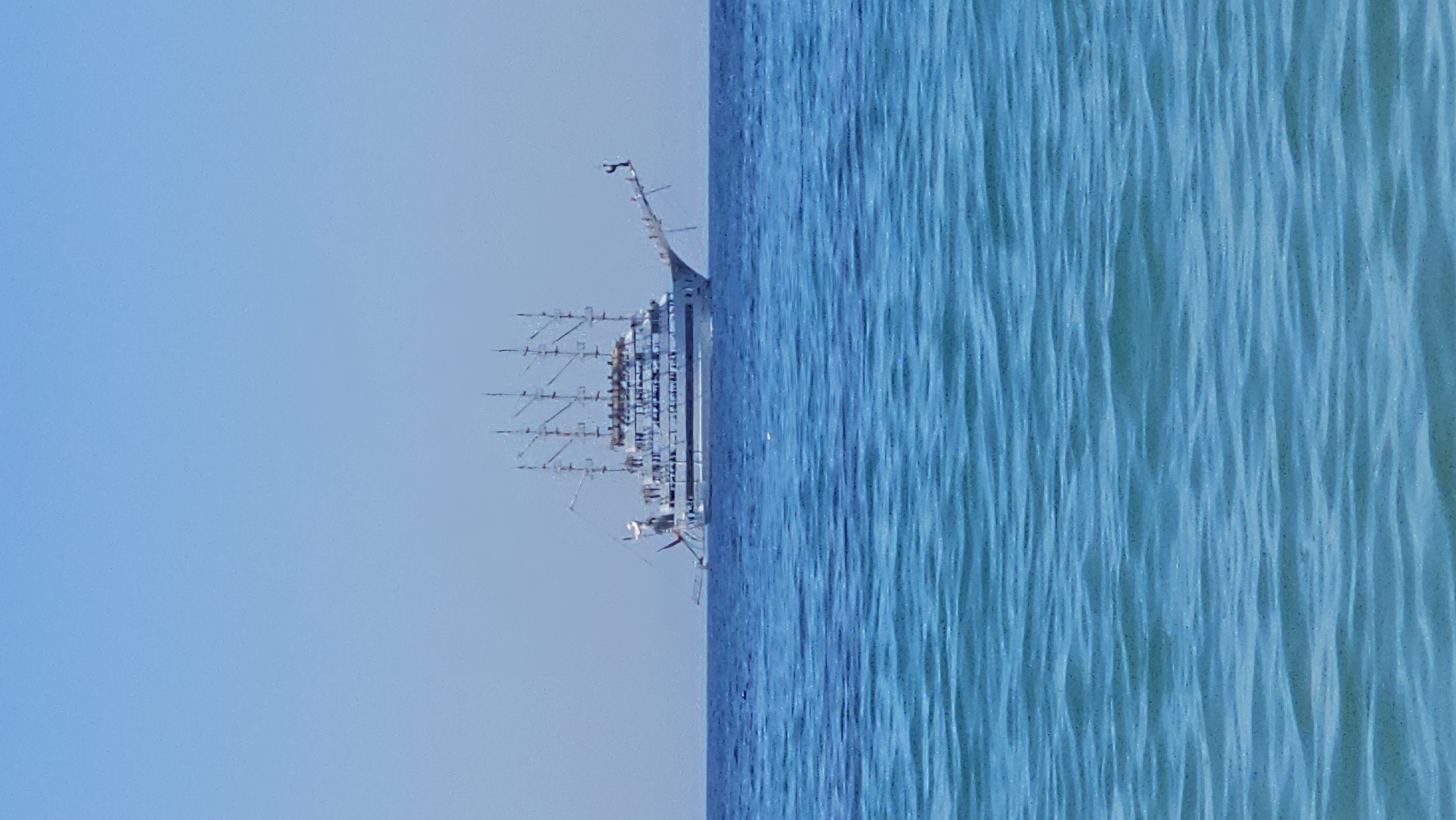 A large white sailing ship with multiple masts is navigating the ocean under a clear blue sky. The ship appears to be moving from left to right. The water in the foreground is a vibrant turquoise, becoming a deeper blue as it recedes towards the horizon where the ship is located. The image composition suggests a view from the coast of Alanya, Turkey, looking out towards the sea on a sunny day.