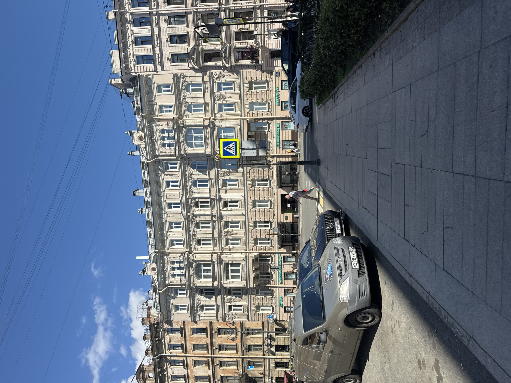 The image depicts a street scene in Saint Petersburg, Russia, on a sunny day. In the foreground, a light gray utility van is parked, partially shadowed. To its right, a dark-colored sedan is parked, and a white sedan is visible further down the street. Pedestrians are walking on the sidewalk, and a crosswalk is present. Several apartment buildings with classical architectural styles line the street, featuring multiple stories, numerous windows, and ornate facades. Overhead, power lines crisscross the sky. The sky is blue with some white clouds. The overall impression is of a typical urban environment in Saint Petersburg.