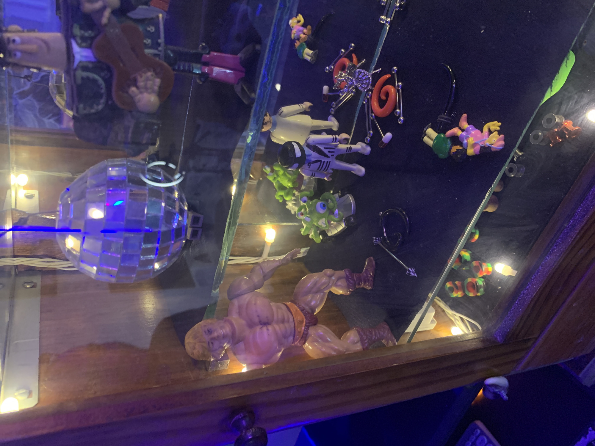 A cluttered display case containing a variety of items. From left to right, there is a disco ball, a figure holding a guitar, several sets of ear stretching jewelry, figurines of various characters, and an action figure of He-Man. The lighting is dim, with multiple lights visible. The environment consists of a wood-framed display case. Location is not deducible from the information in the image.